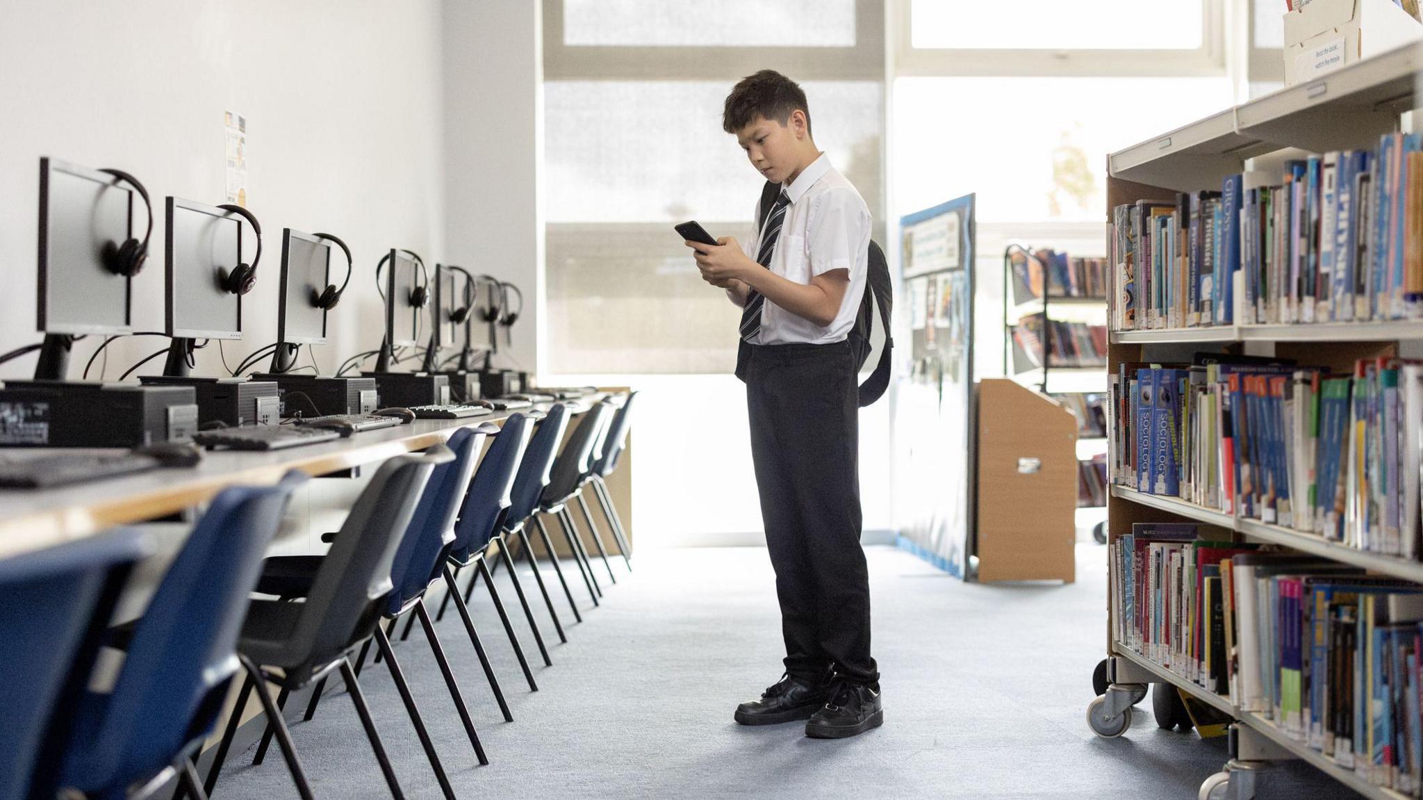 A boy in school uniform stands in the library looking at his phone