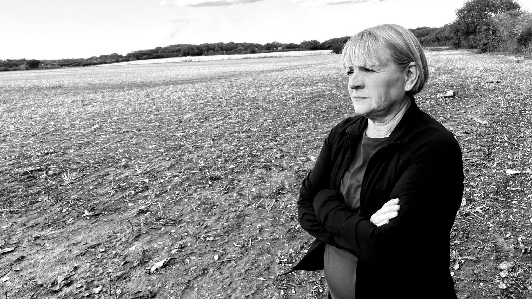 Vanessa Jones stands looking across a field, looking concerned, and with her arms folded in this black and white photo. She is wearing a dark coloured top and jacket. Shrubs and trees can be seen in the distance.