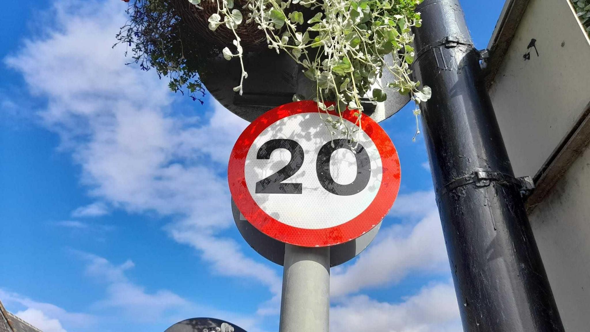 A 20 mile per hour road sign pictured against a blue sky background. The sign is white with a red rim and the number 20 is in black. Three is a black lamppost next to the sign and it is held up on a grey post.