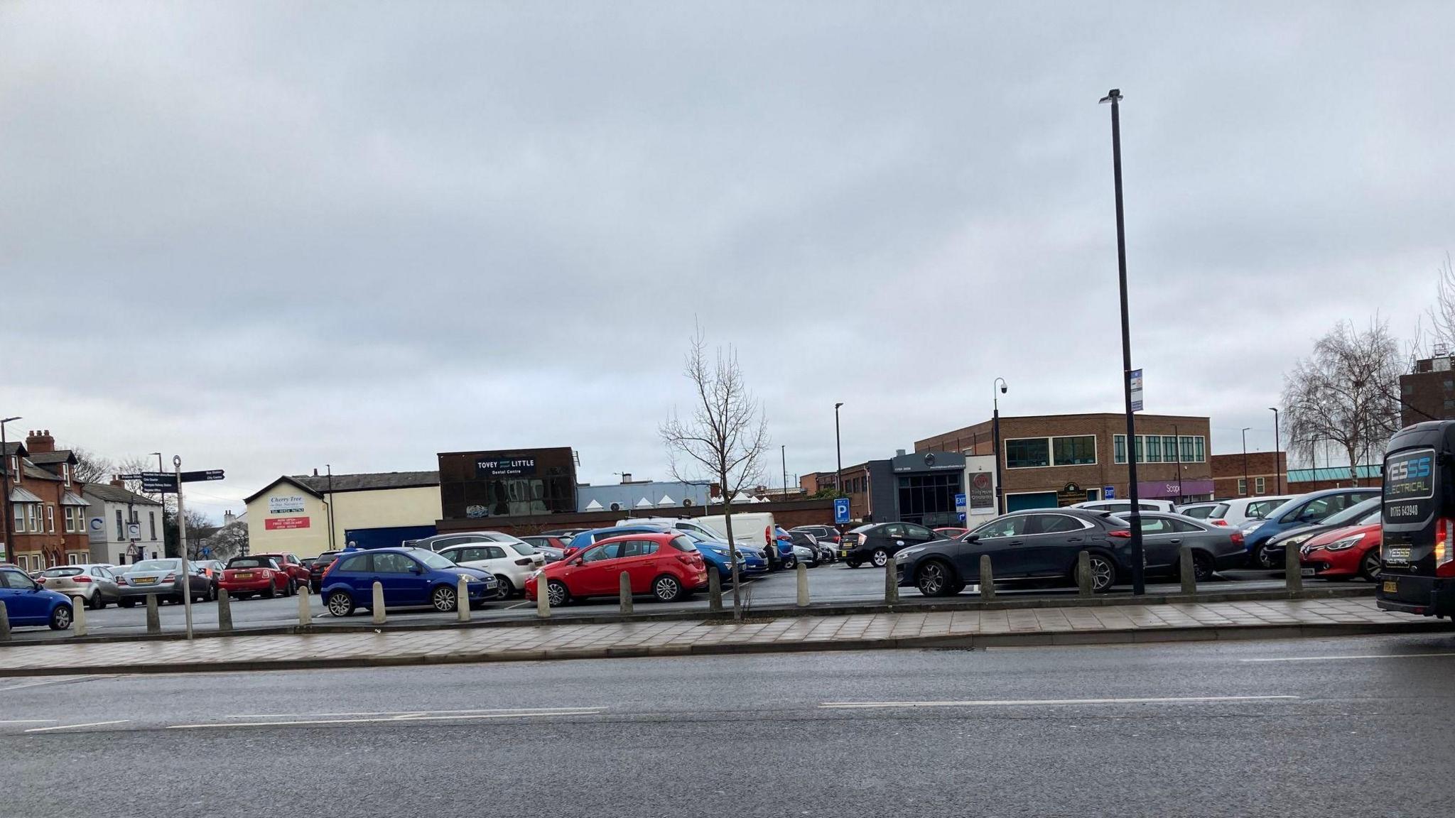 Picture of Borough Road Car Park in Wakefield City centre. It shows a number of cars parked, with a road in the foreground and buildings in the background. 