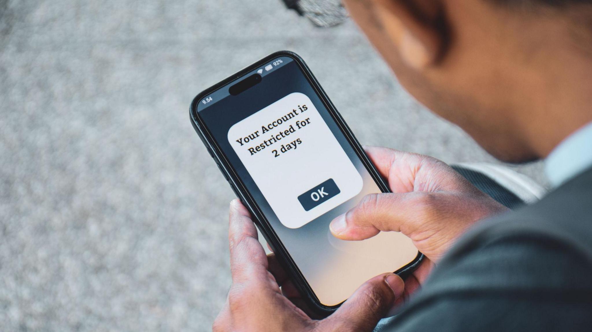 Person with brown skin holding a mobile phone which has the message "Your account is restricted for 2 days"