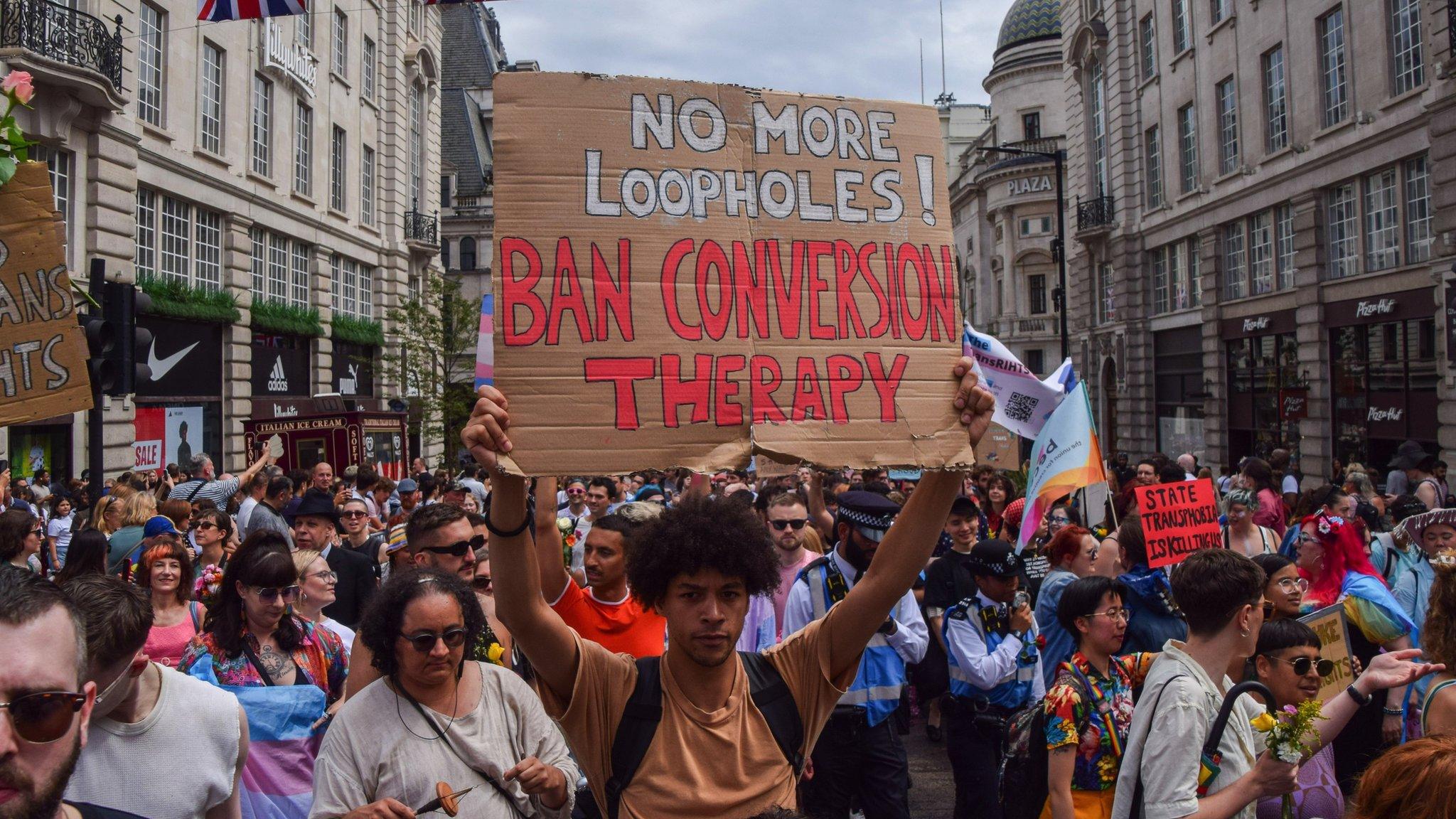 A man in a crowd holds up a sign that says "No more loopholes! Ban conversion therapy"