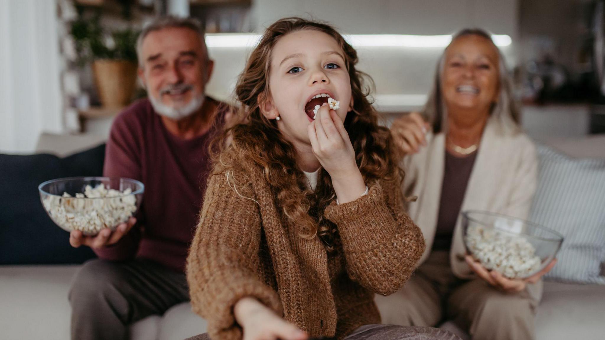 Grandparents watching TV with their granddaughter and eating popcorn