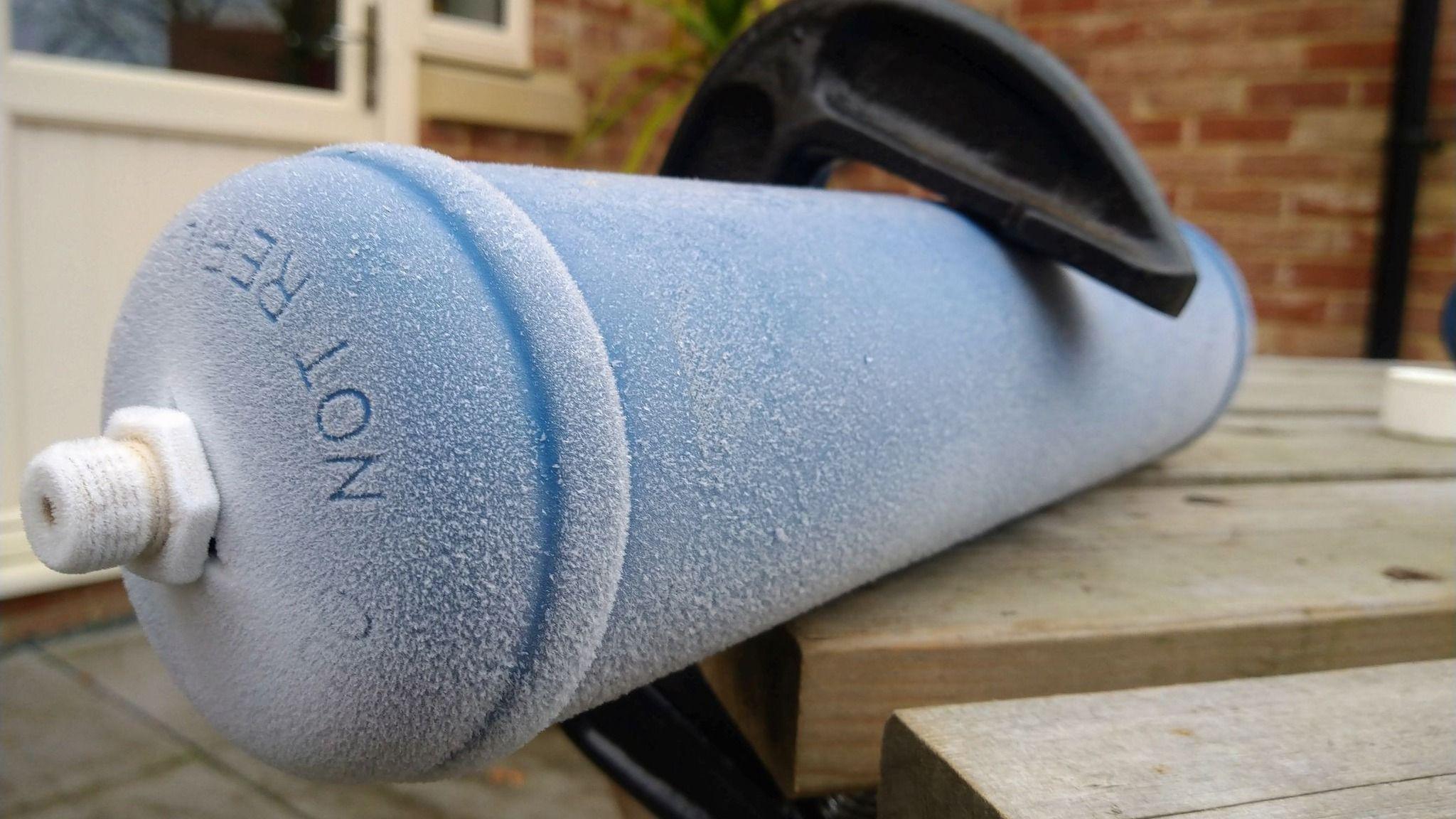 A blue canister attached to a garden bench with a metal 'G' clamp. The can contained a small amount of nitrous oxide, which when released froze the canister over in a thin film of frost. The brickwork, door and window of a home can be seen behind the canister.