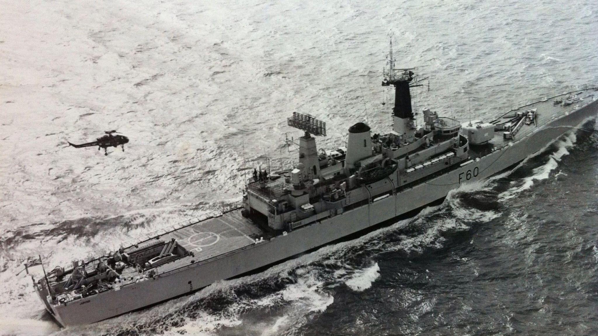 An aerial shot of HMS Jupiter. The black and white image shows a long, slim battleship at sea, with a helicopter flying over it.