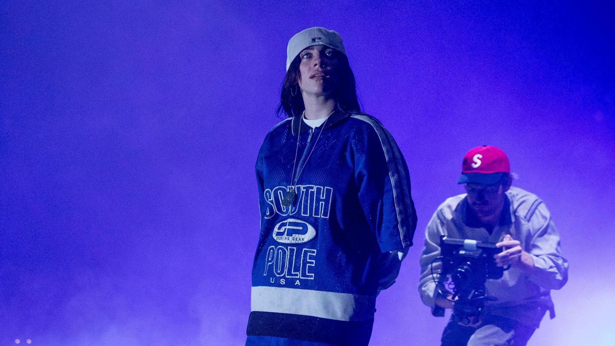 Billie Eilish performing on stage. Billie wears an oversized blue jumper and a backwards twisted white baseball cap. She has long dark hair and is lit with blue lights. 