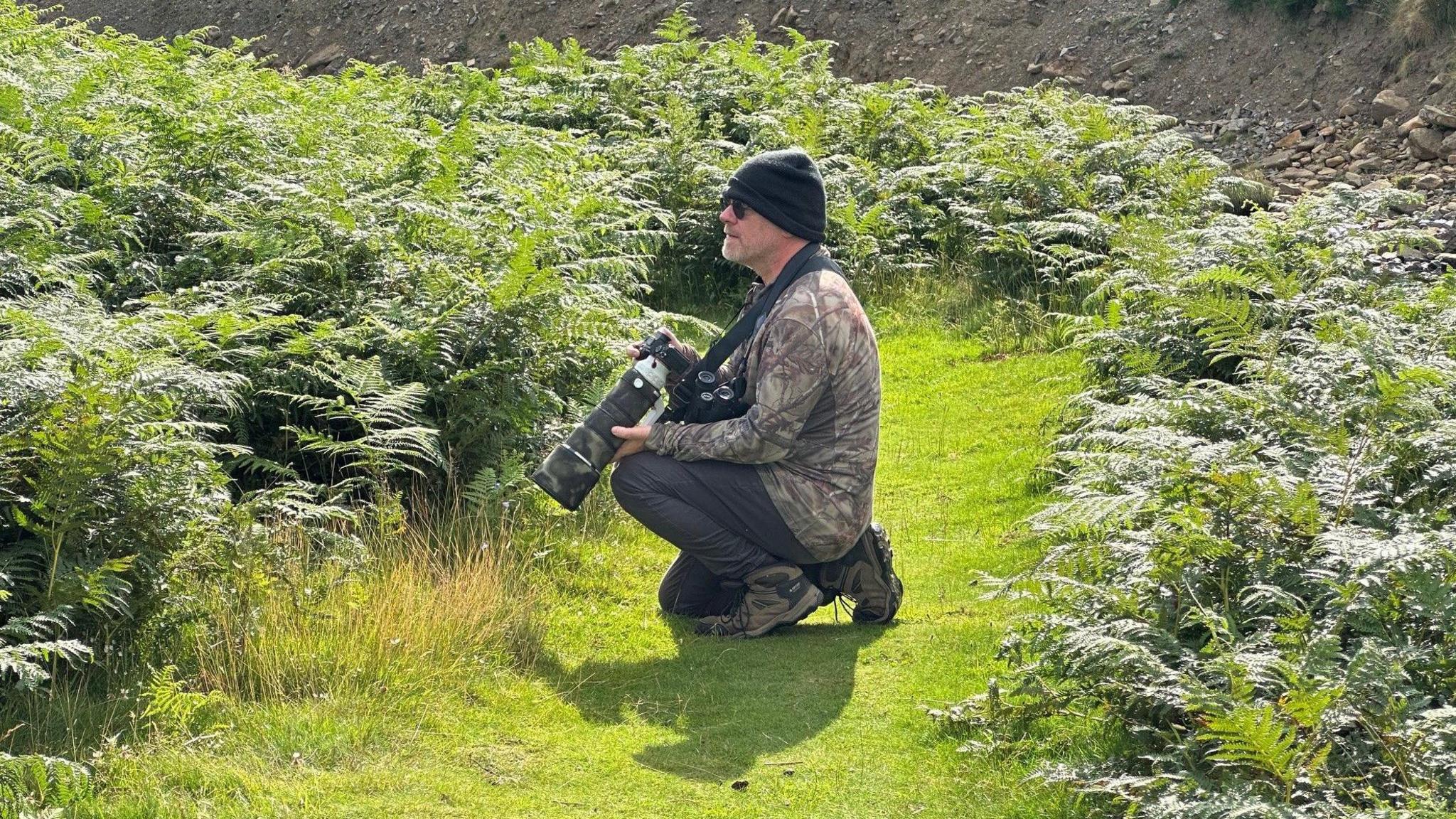 A man in a camouflage jacket crouches next to some ferns while holding a large camera in his hands.