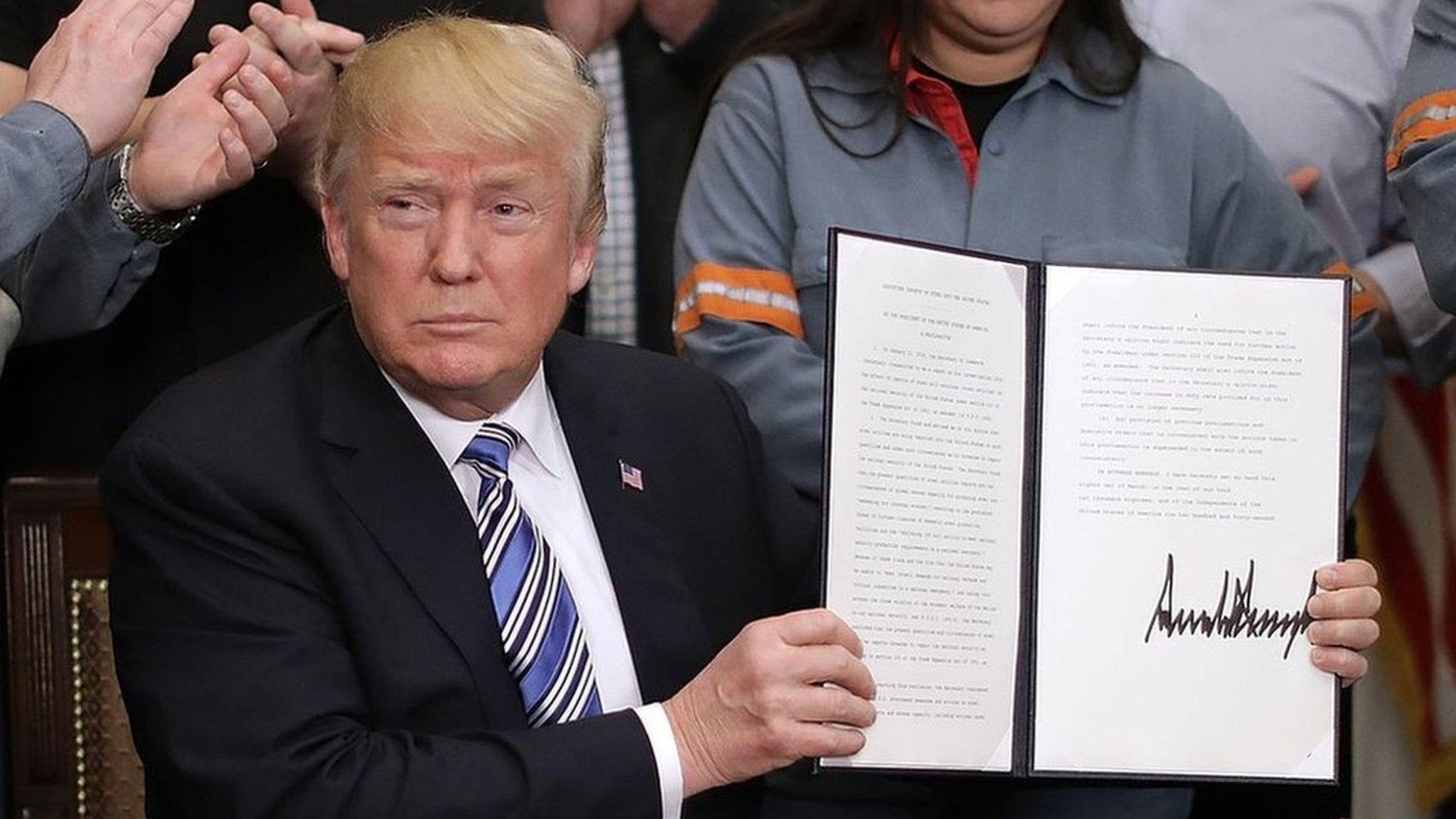 President Donald Trump holds up a piece of paper with his signature while surrounded by steel workers in uniform