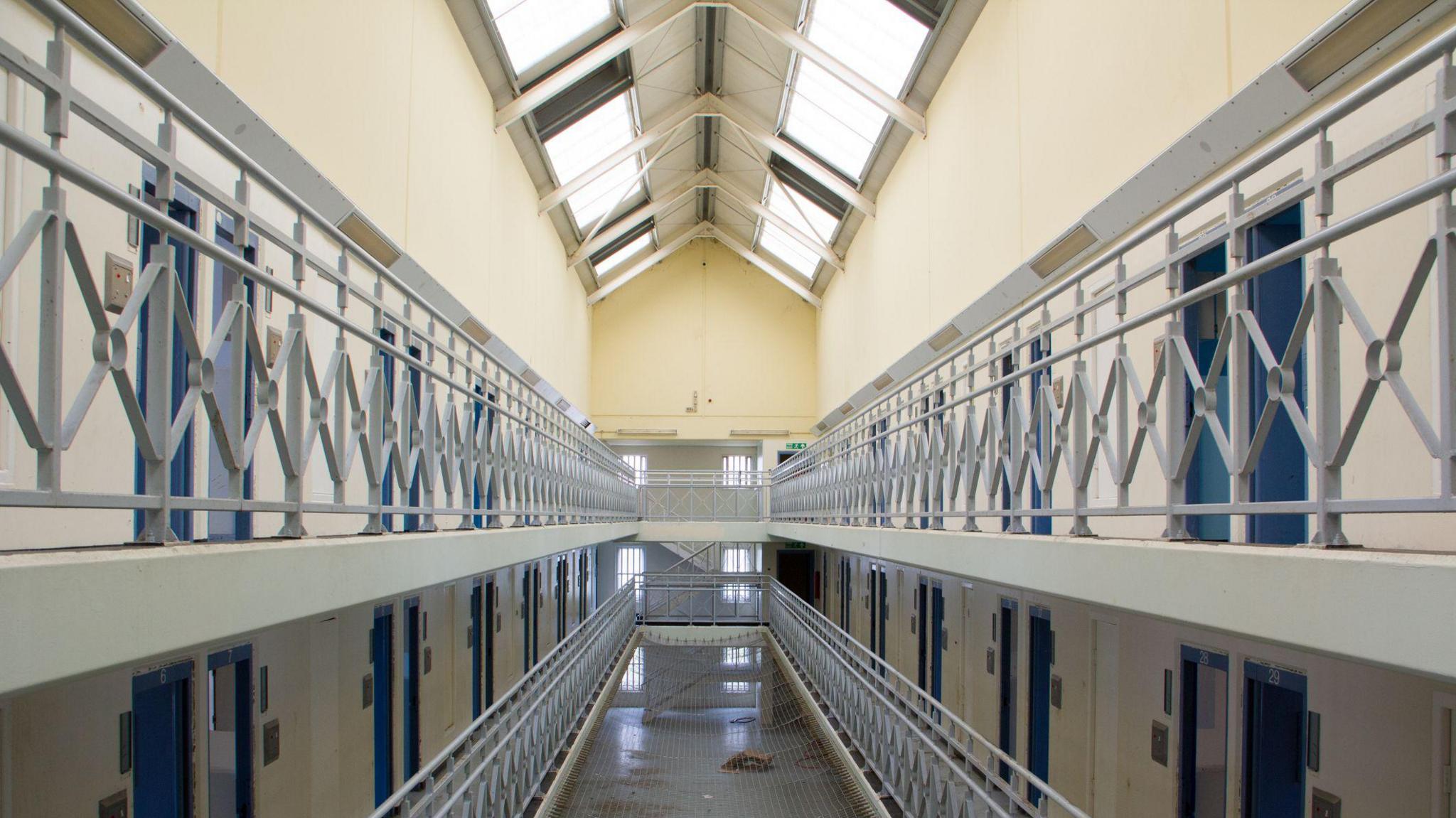 Empty corridors in prison with yellow walls and lots of doors open. Building has lots of natural light and is multi-storey. 