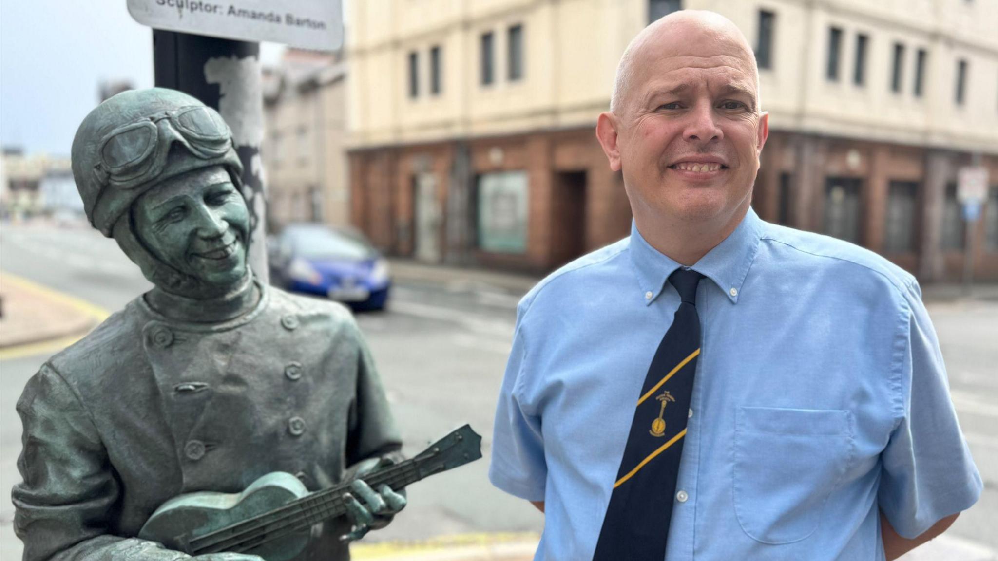 Andrew Poppleton wears a blue shirt and a navy tie, he has short grey hair. He stands next to a green statue of George Formby who wears a helmet and goggles and plays a ukelele.