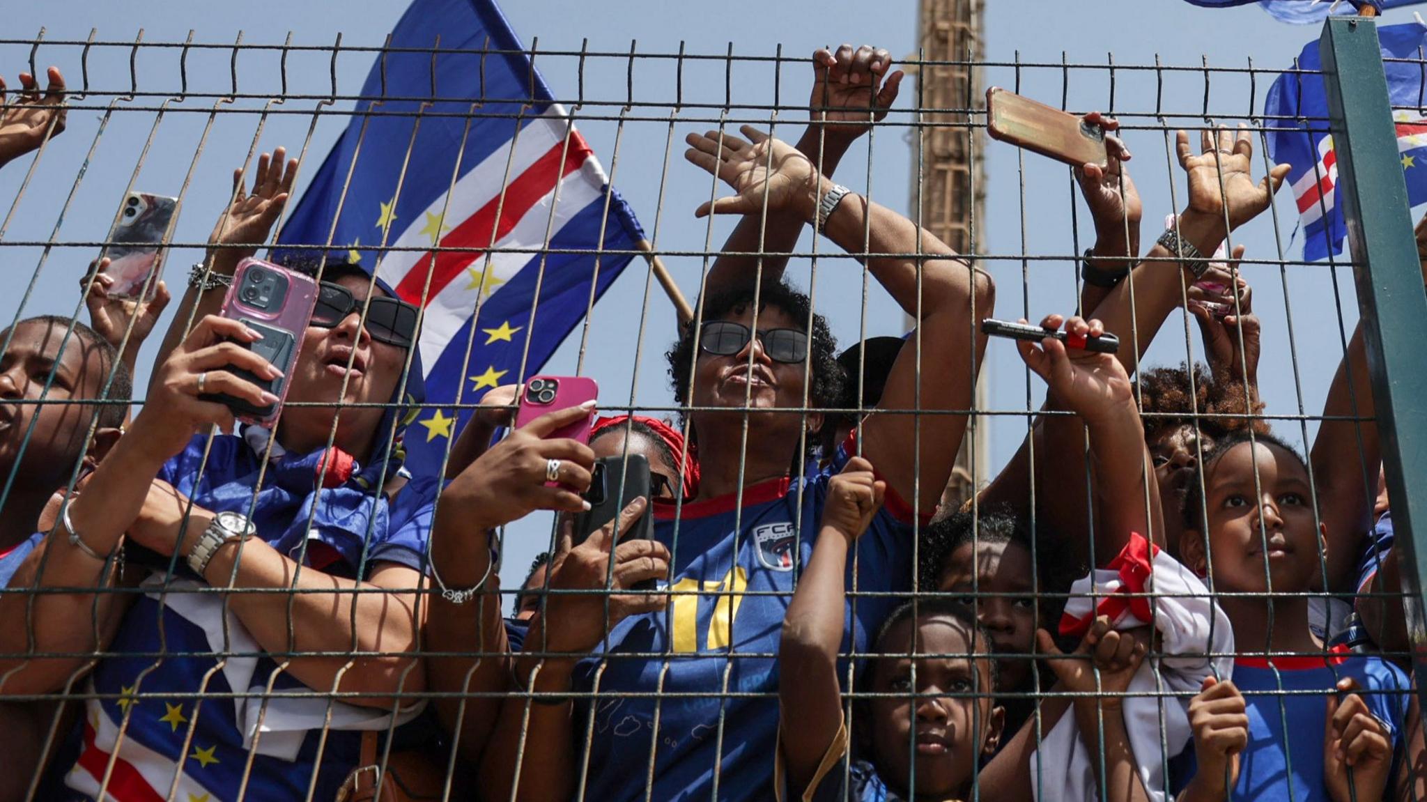 Fans wearing the Cape Verde kit press against a metal fence.