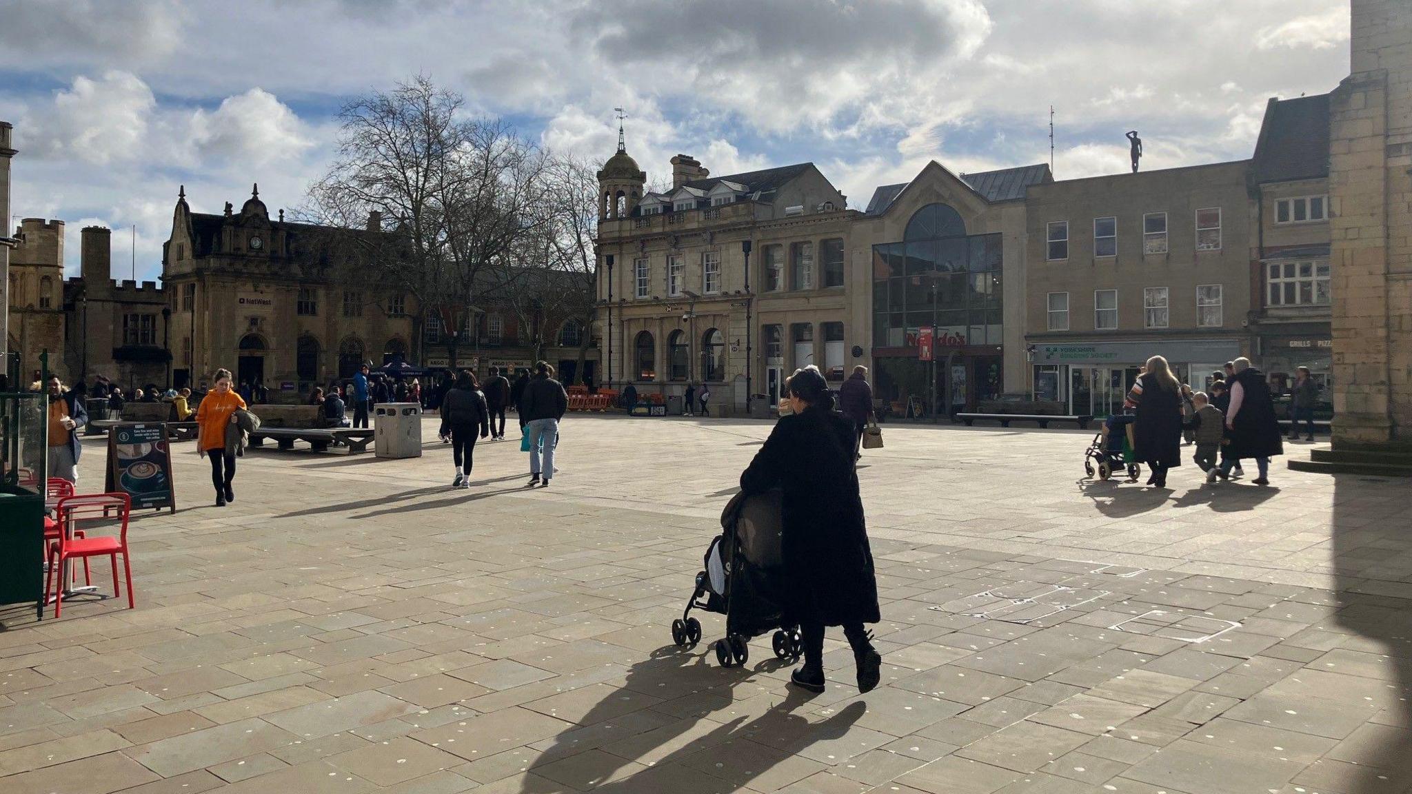 Peterborough's Cathedral Square, including buildings with historic architecture and members of the public walking around.