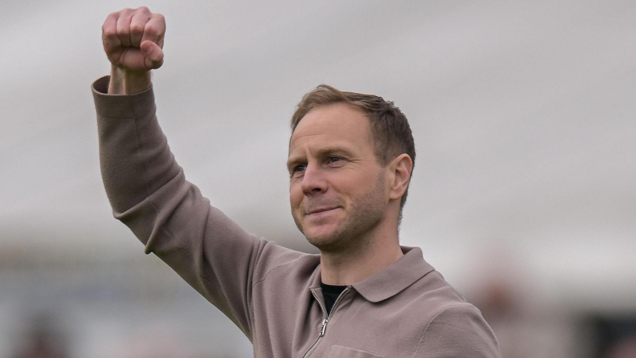 Ian Watson, wearing a beige zip-up jumper, has his right fist clenched and held up in celebration after South Shields' win over Buxton