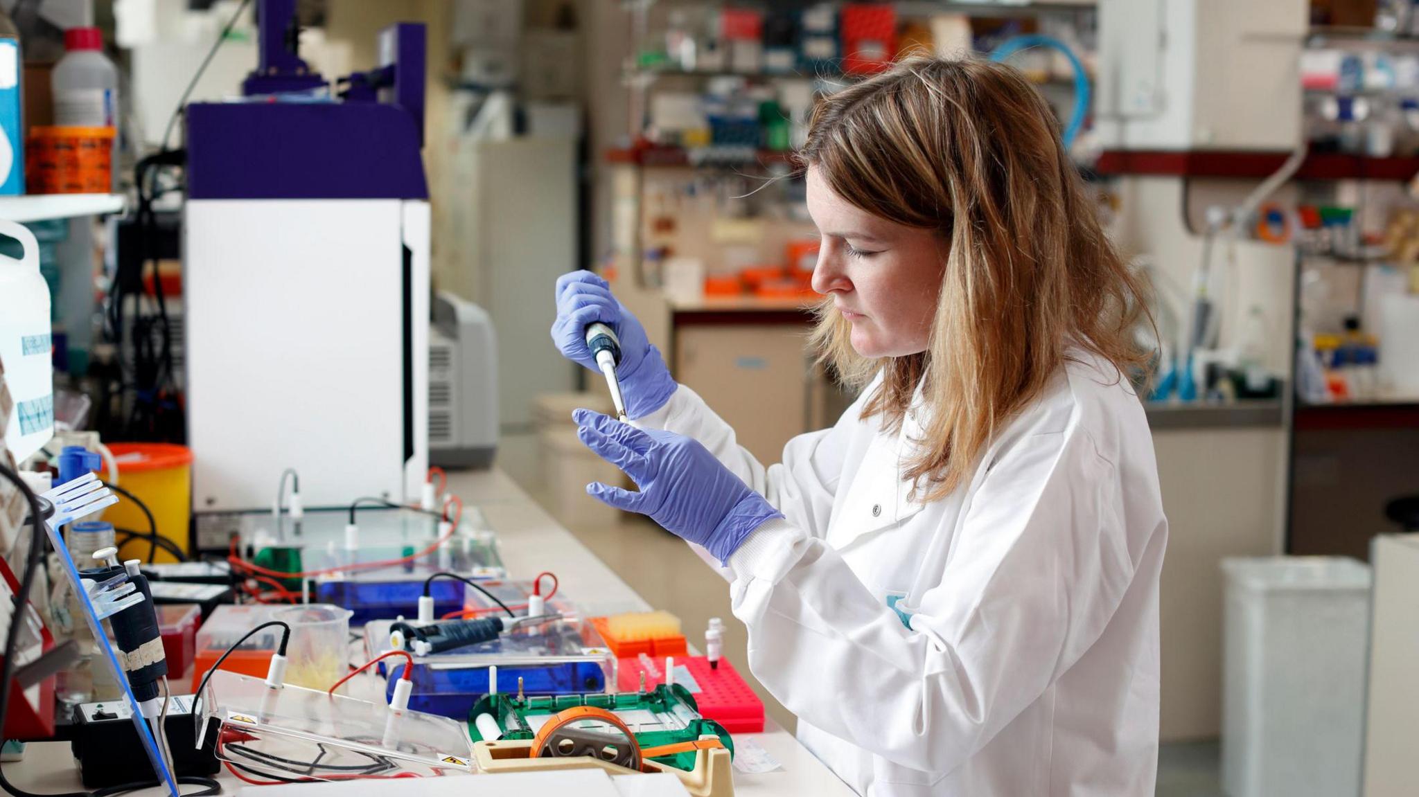 A woman in a laboratory wearing blue medical gloves and a white lab coat administers a solution in a vial using a pipette.