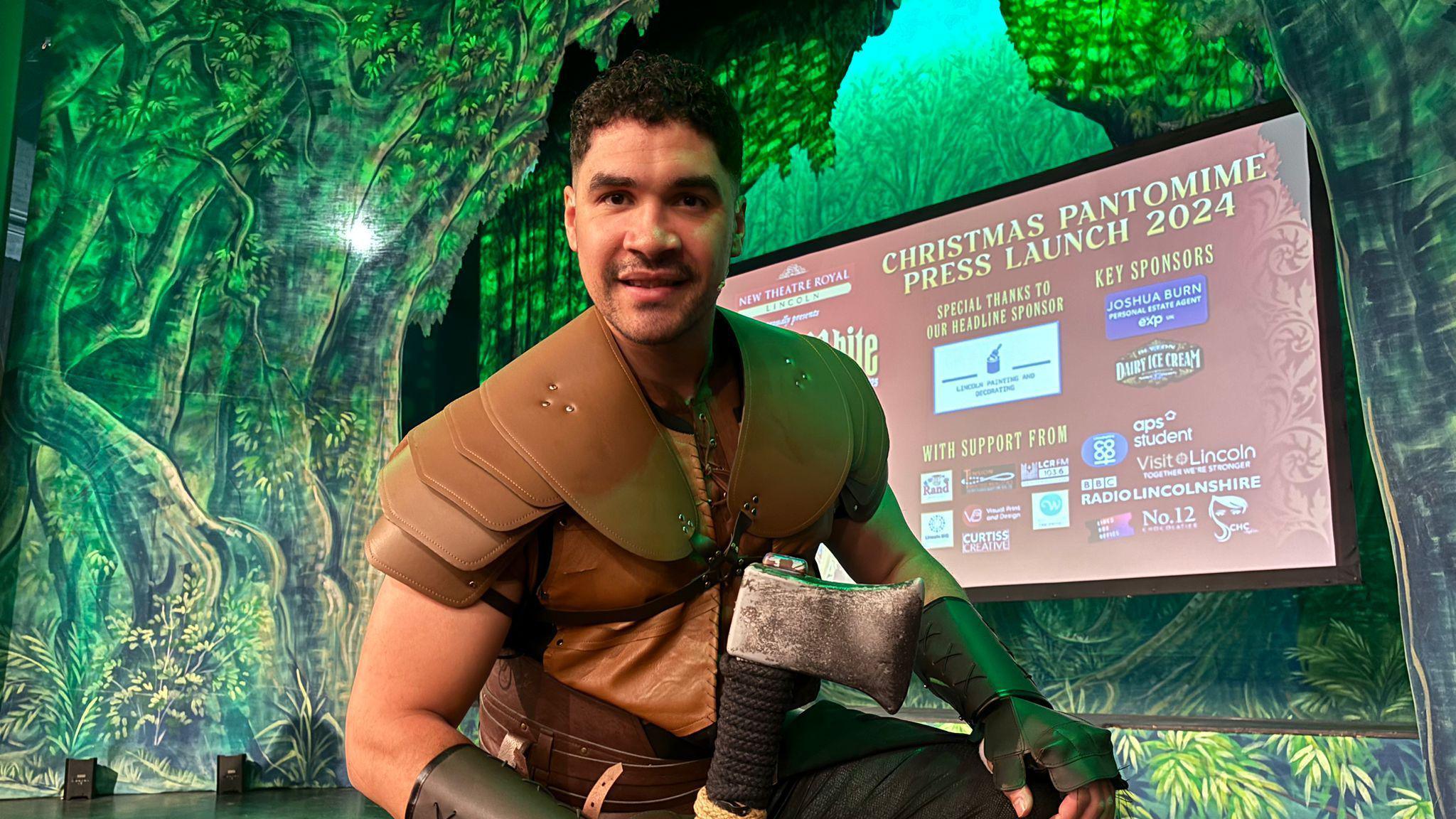 Olympic gymnast Louis Smith in a huntsman costume on stage at a theatre. The backdrop shows green foliage.