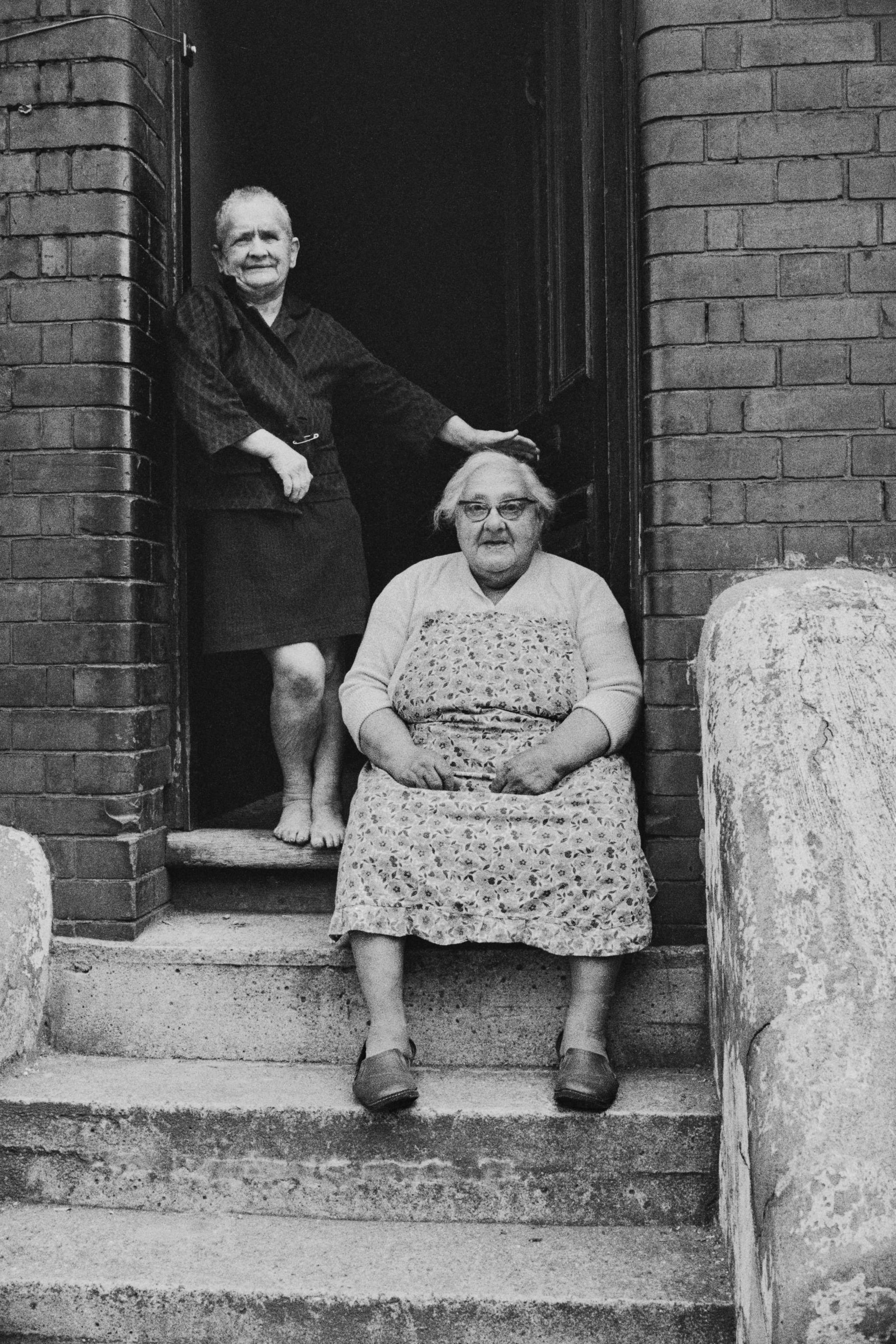 A black and white image of an elderly woman sitting on some steps in front of a building with her daughter standing in the doorway behind her. 