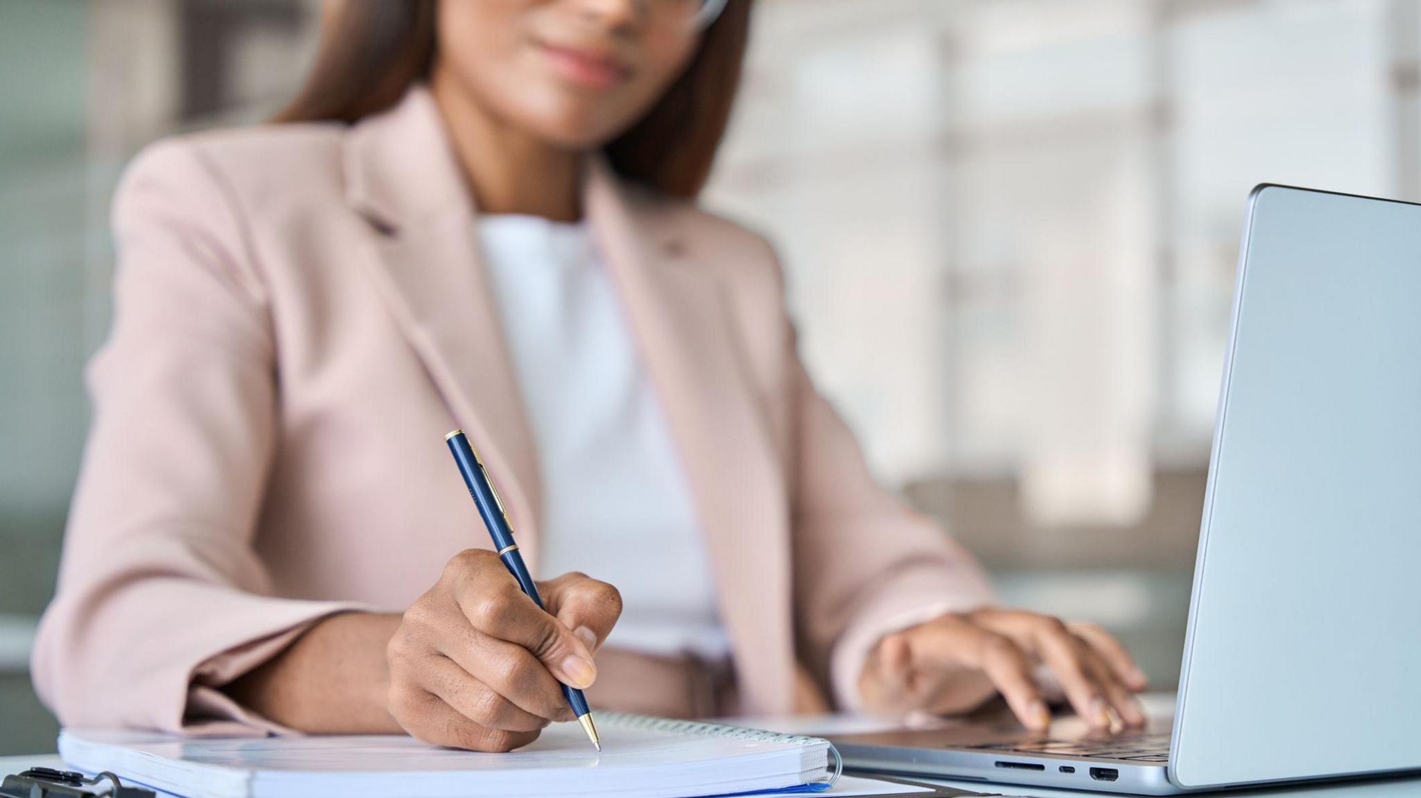 A woman, whose face is only half visible and slightly out of focus, wearing a pale pink jacket, writes using a smart ballpoint pen on a spiral bound notebook. Her other hand is using the keyboard of a laptop that is open on the table in front of her.