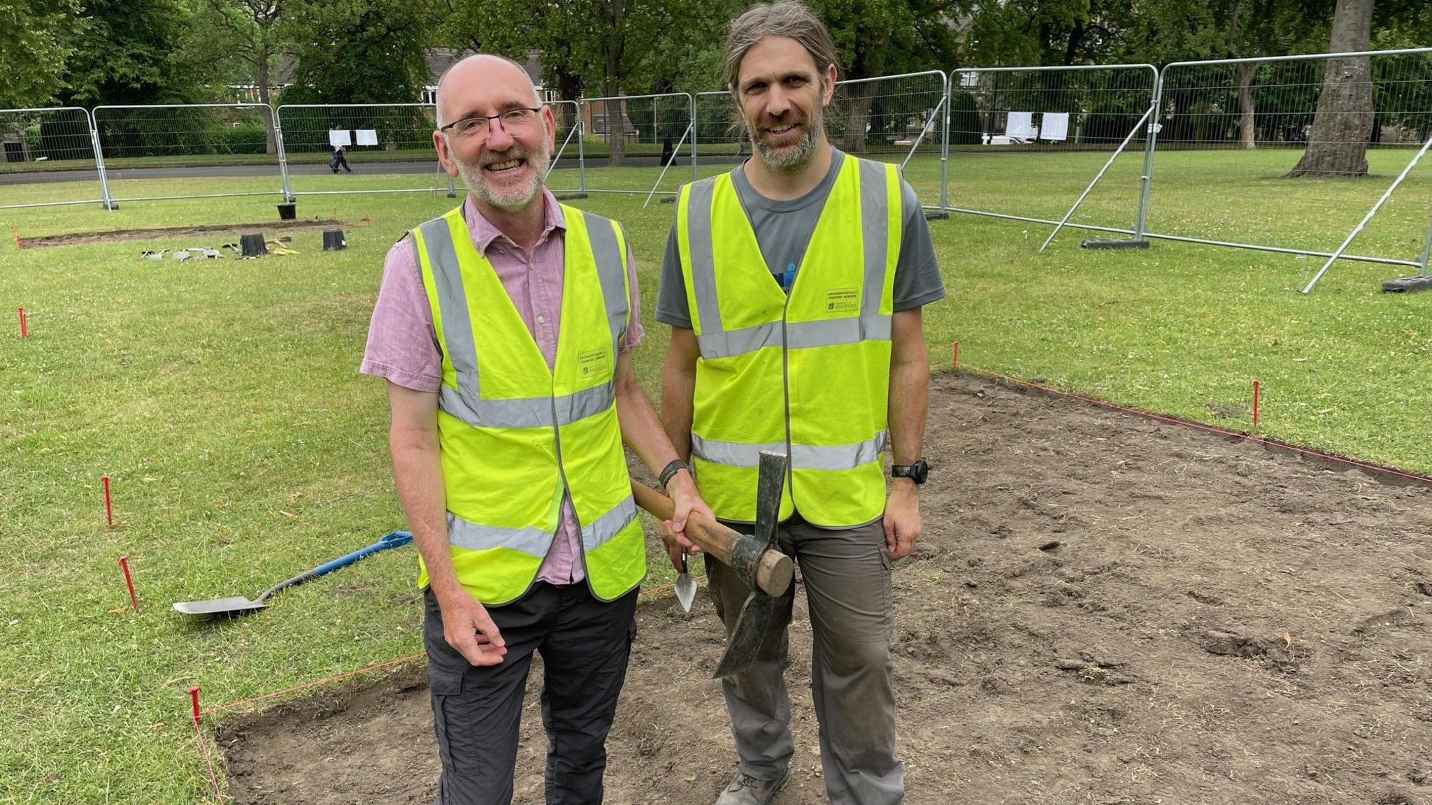 Two men wearing high visibility yellow jackets stand in a dug up part of a field which is fenced off. They both carry digging tools and are surrounded by others on the ground.