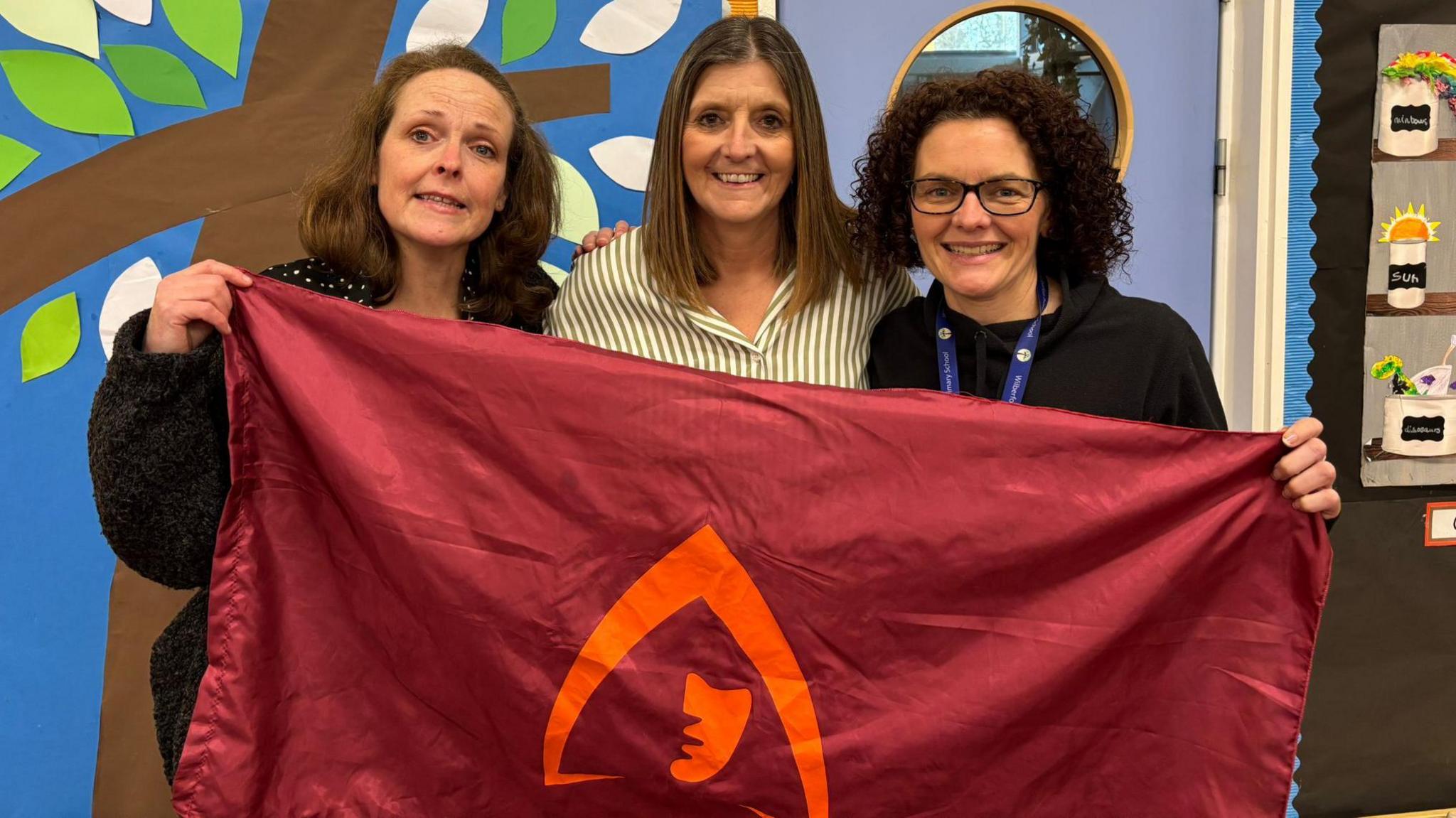 Kirsten Cooper (left), Jo Dyer (middle) and Mel Cartmail (right). They are all holding a red flag with an orange logo in the centre. 