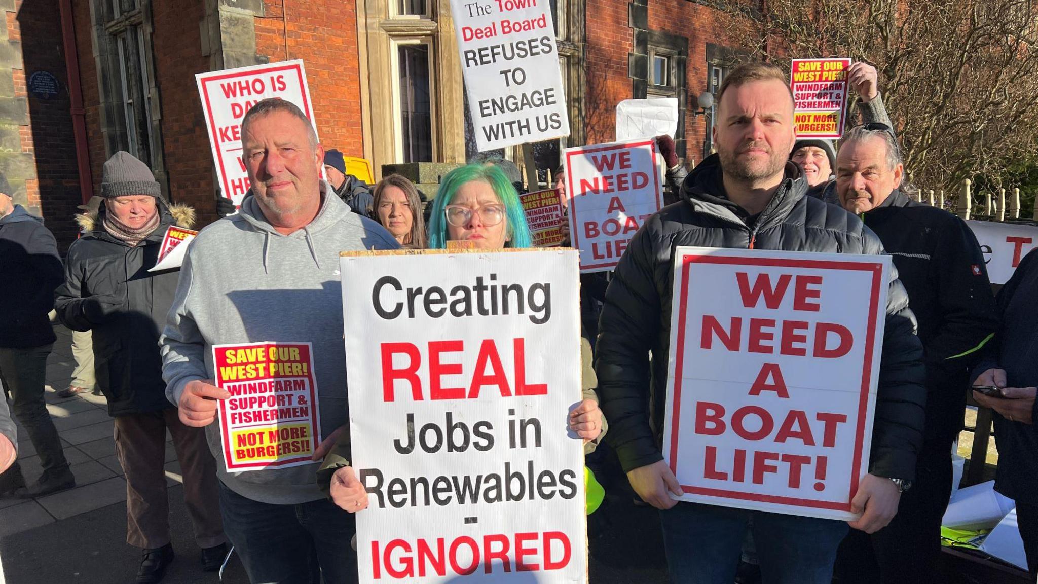 Men and women stand with placards that read "creating real jobs in renewables - ignored", "we need a boat lift" and "save our West Pier, windfarm support and fishermen, not more burgers".