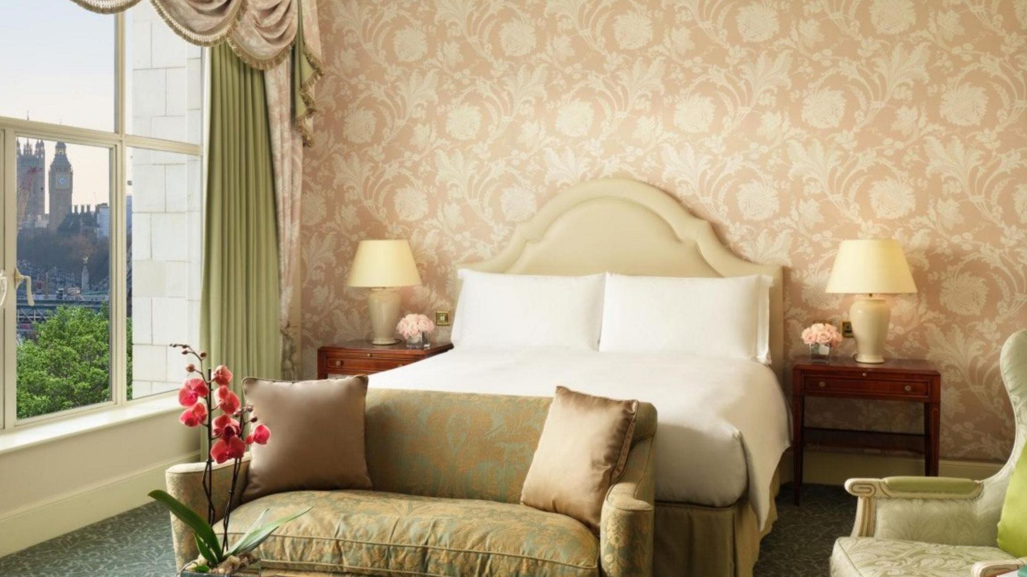 A room in The Savoy with a cream bed with white sheets, a green patterned sofa, wooden bedside tables and pink and green curtains and drapes.