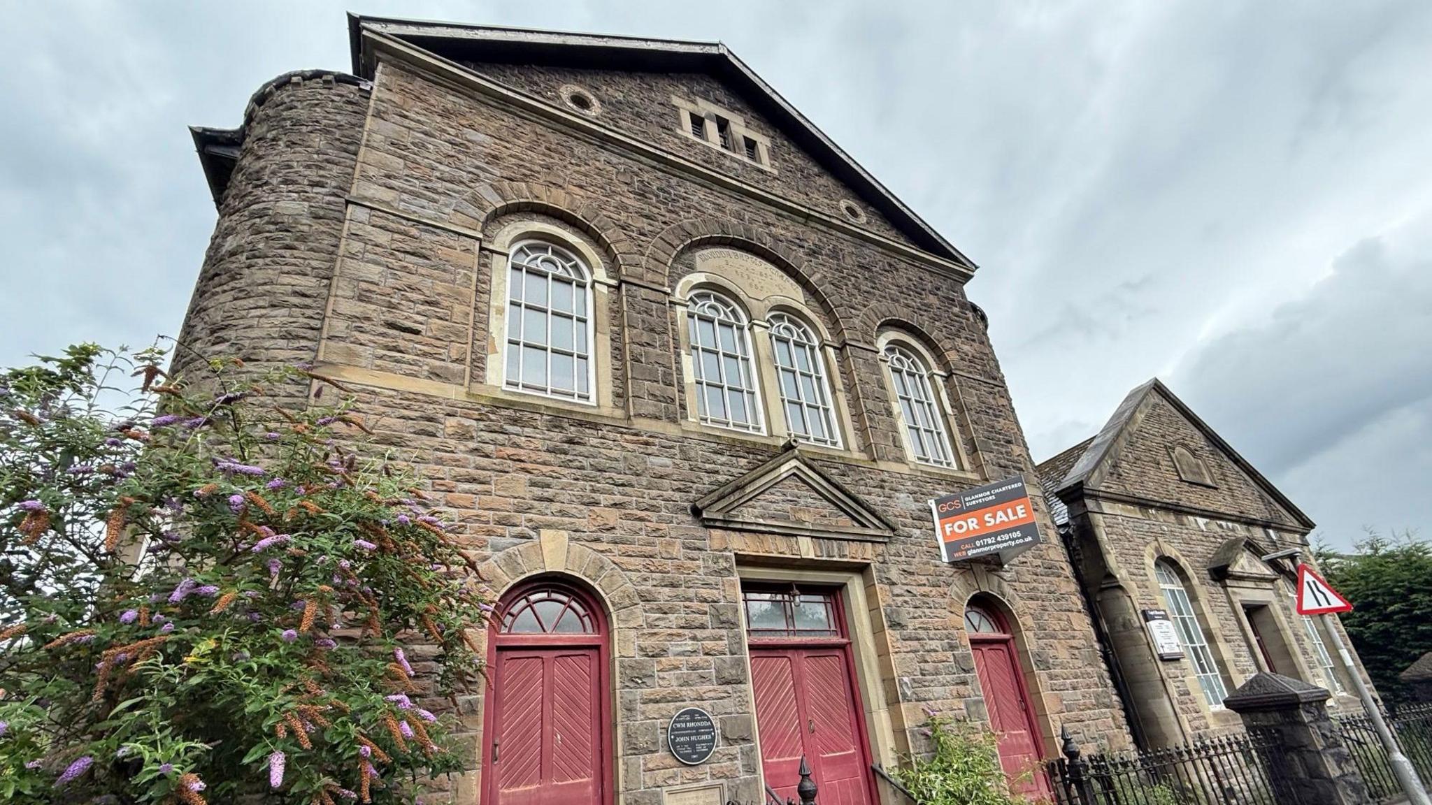 Front façade of the chapel with three double wooden doors side by side painted dark red and four arched windows above. An orange for sale sign has been attached to the wall at the front.