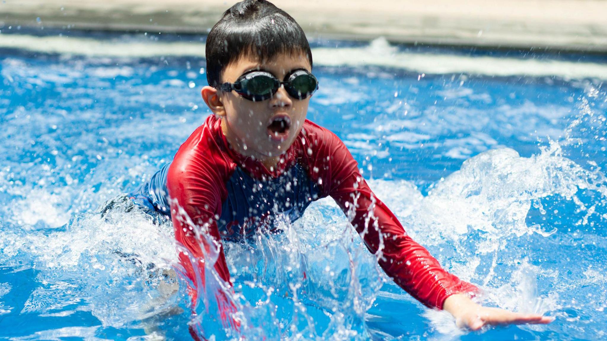 A young boy wearing a red wet suit and black goggles splashes in a swimming pool.