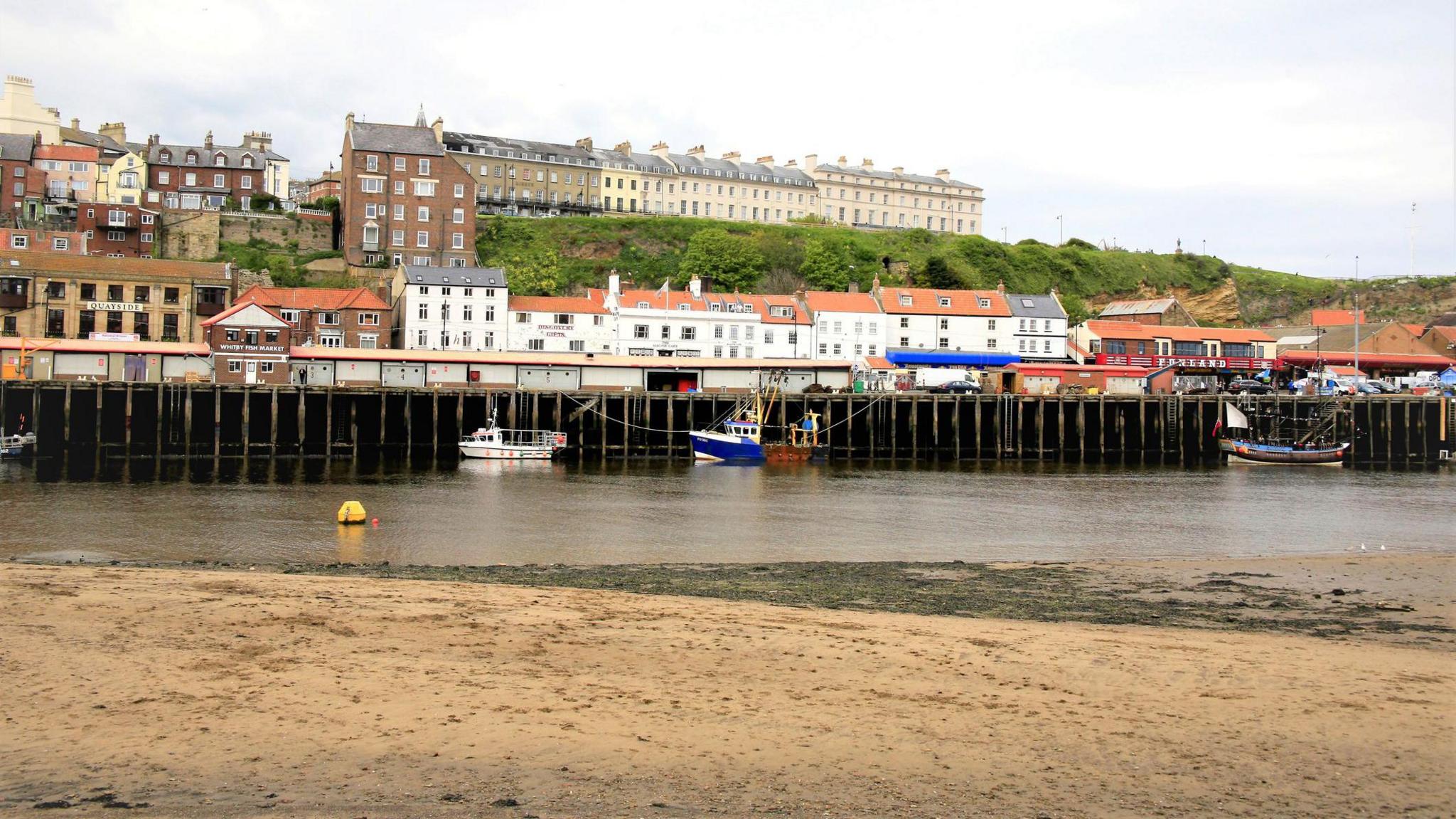 Whitby Fish Quay. The picture is taken from a distance. The oak fenders, which will be replaced as a result of the work, can be seen. Three boats can be seen alongside the quay. Some of the red-roofed buildings of Whitby can be seen in the distance. 