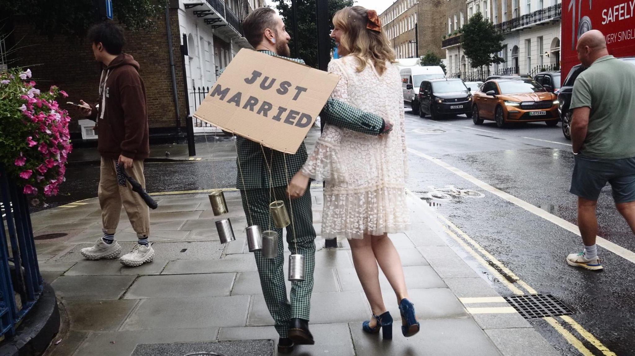 Chris and Sam Jamieson-Green walk along the street. Sam is wearing a white wedding dress and has her arm around the back of her new husband Chris who is wearing a green and white chequered suit with a cardboard sign saying 'just married' on his back.