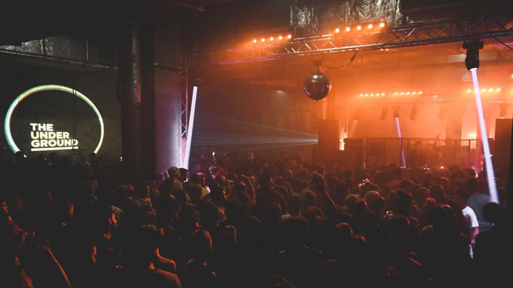 A big crowd of people are seen dancing in a dimly-lit room under The Galleries in Bristol. There is a disco ball, and strobe lights.