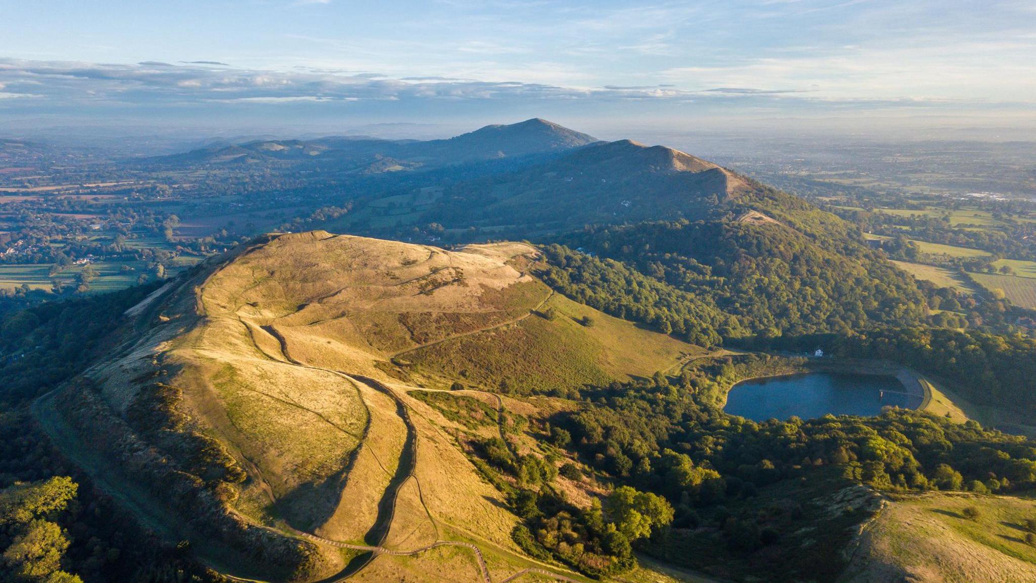 An aerial view of the Malvern Hills, they are partially covered in trees and a large body of water is visible