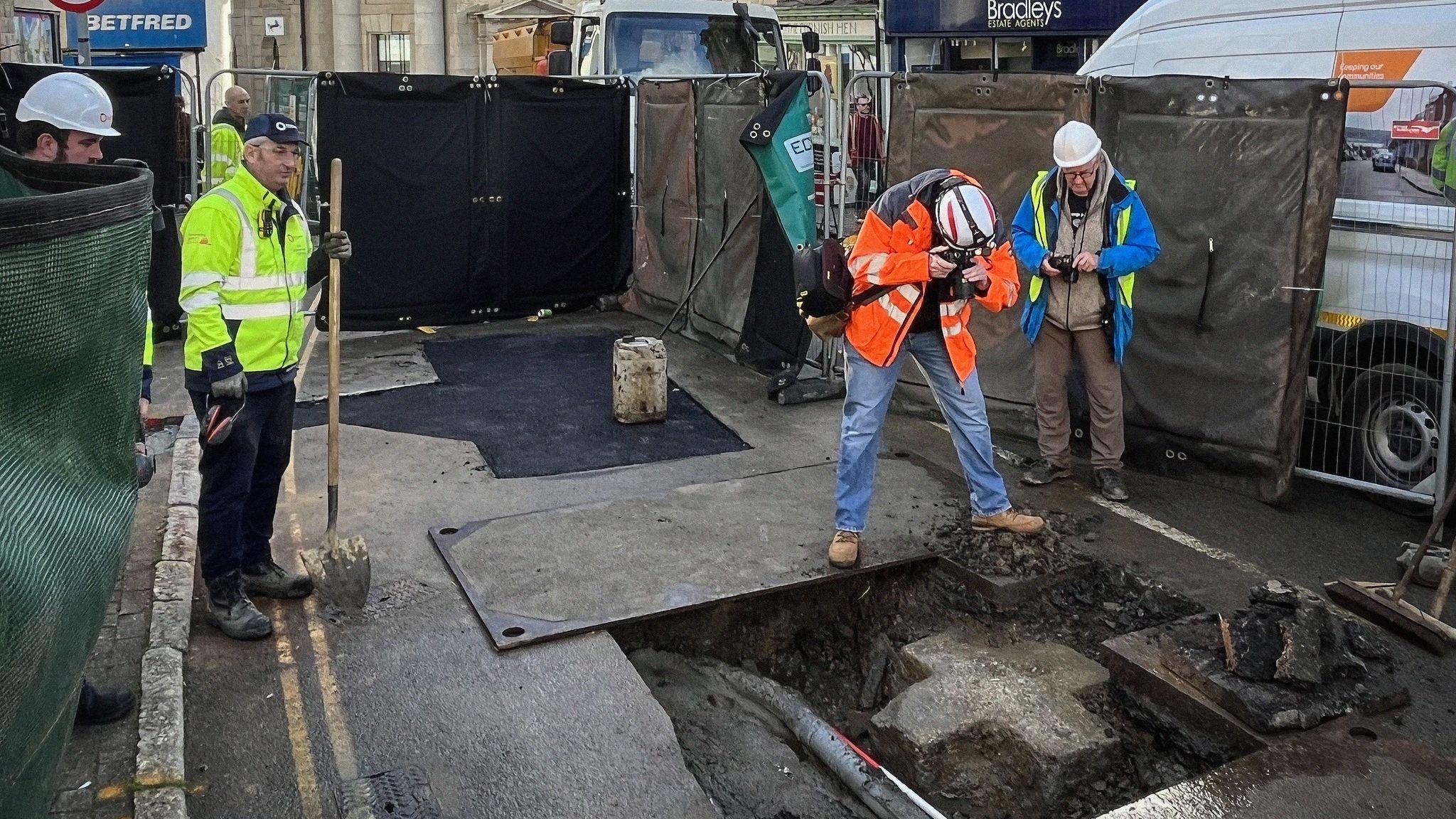 The picture shows a street excavation site. The scene includes a large open trench in the road surface, exposing pipes and a section of granite beneath the tarmac. In the trench is a large equal-sided cross. Two workers in high-visibility jackets and hard hats are closely inspecting and photographing the exposed area, while others stand nearby with tools. Safety barriers and fencing surround the site, and construction equipment and vehicles are visible in the background along with nearby shops. 