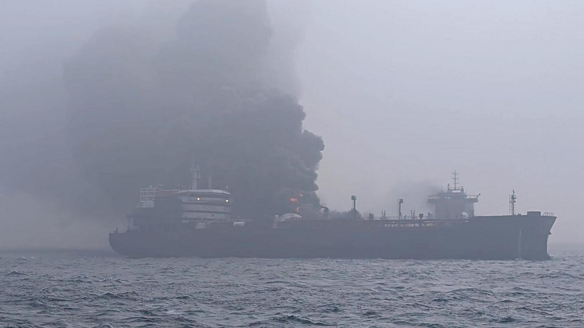 Black smoke pours from two large collided ships with some flames visible. In the  foreground is dark water. Some lights are on in the bridge of one of the ships. The whole scene is slightly silhouetted due to smoke in the air.
