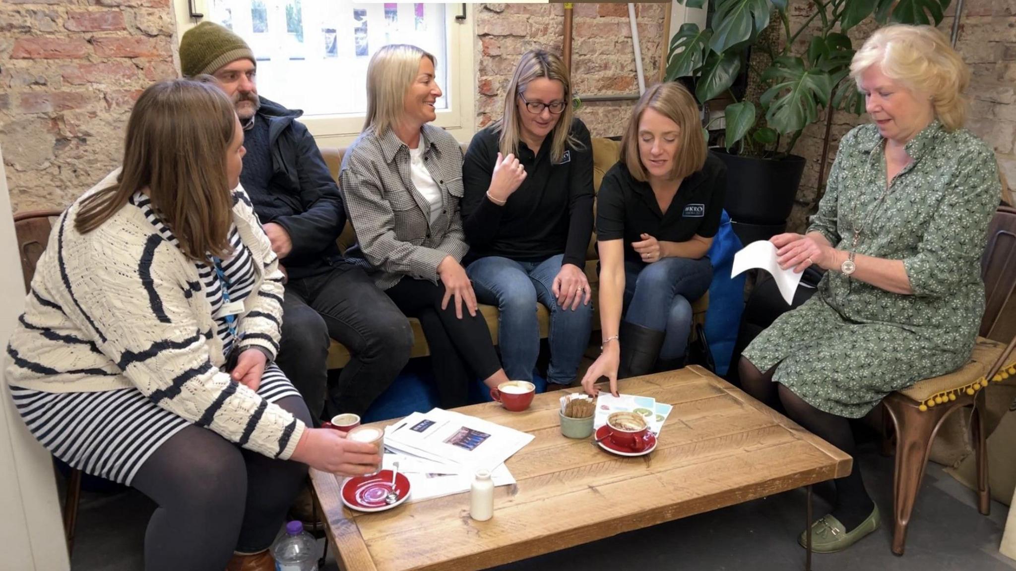 Six people sat around a coffee table chatting and drinking coffee