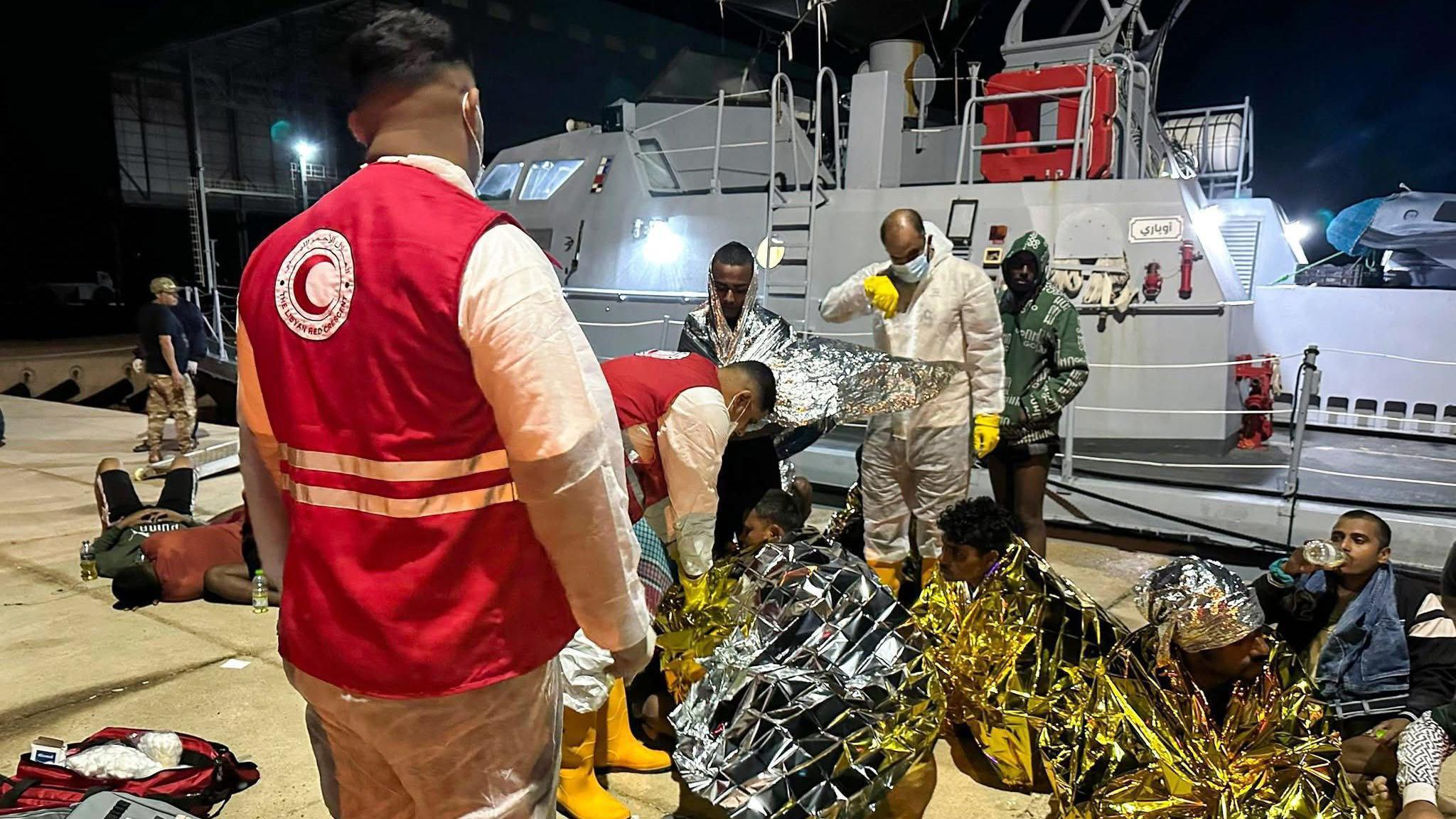 Men in red vests with the Red Crescent logo tending to people wrapped in thermal blankets sitting on the ground. Behind them is a white boat.