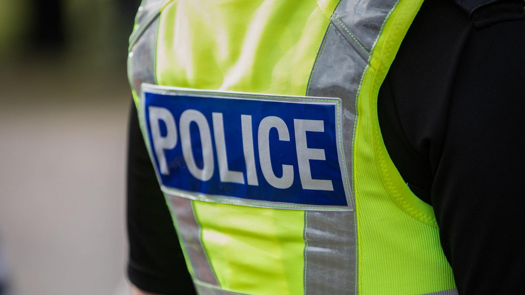 Image of a policeman taken from the back. He is wearing a yellow high visibility vest with the words "Police" in white letters against a blue background 