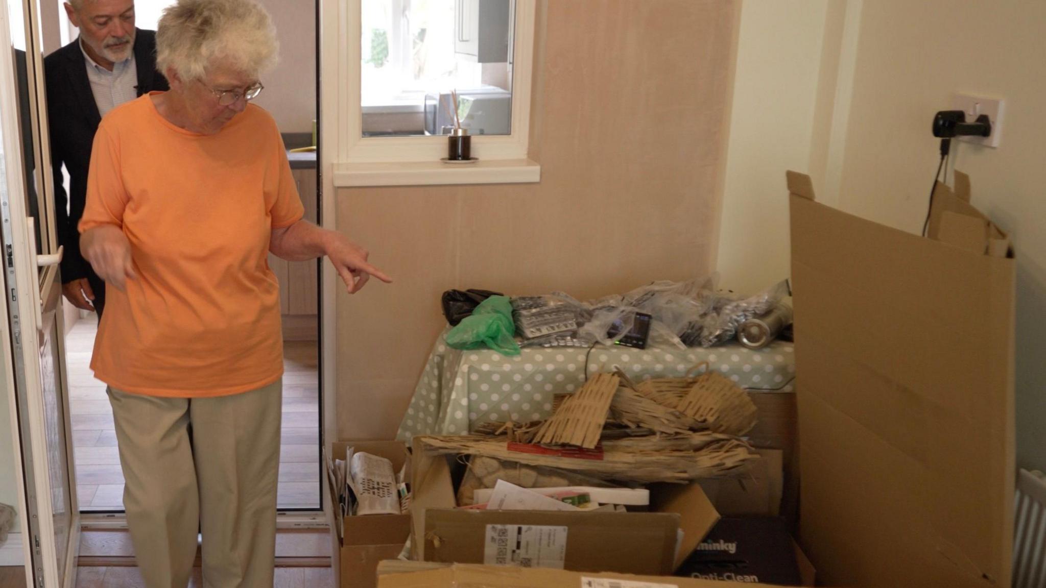 A woman in an orange T-shirt and beige trousers looking down and pointing towards a pile of folded cardboard and boxes of papers and tablet blister packs in a corner of her home.