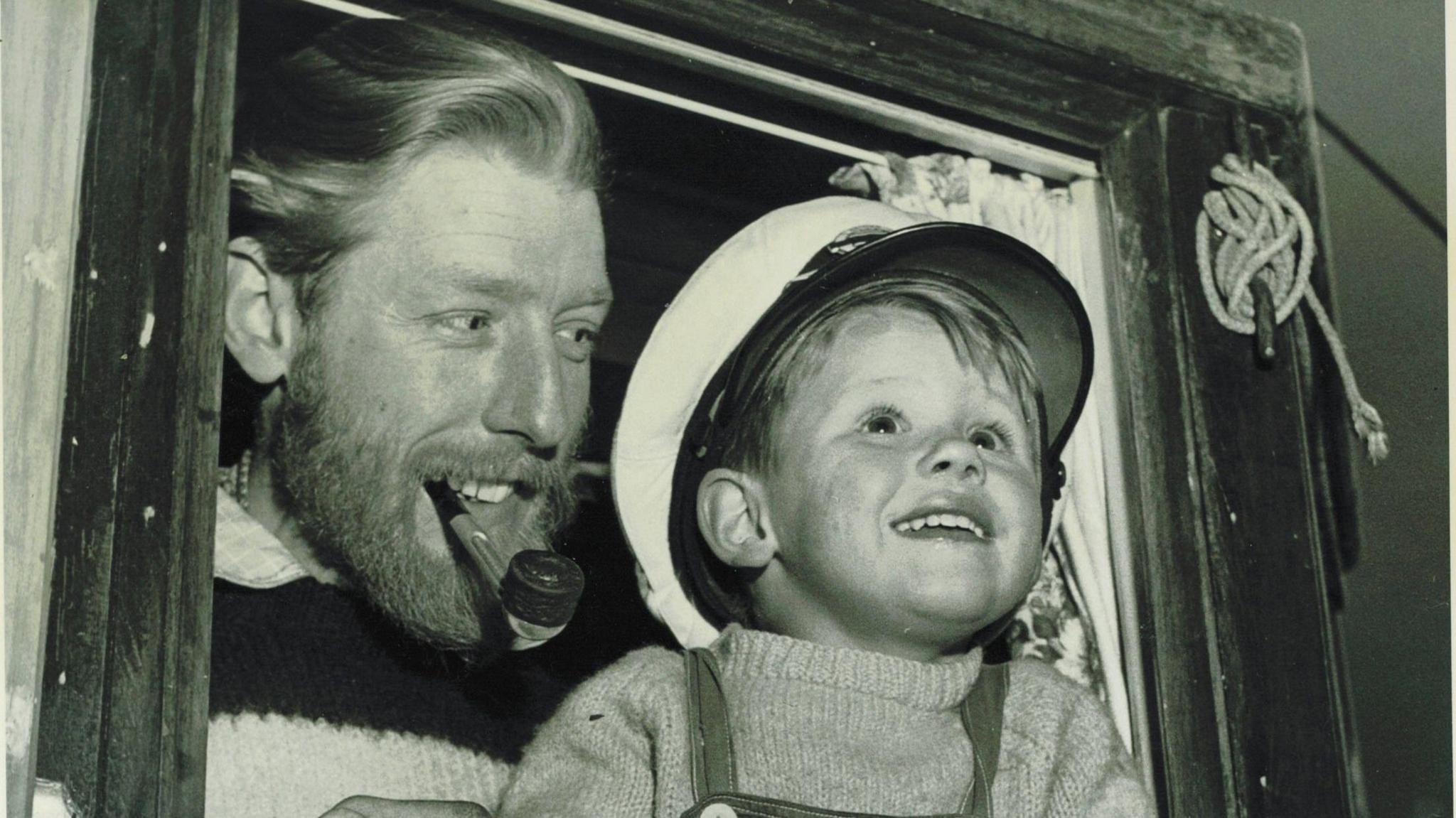 A black and white picture of a young Paul Ainsworth wearing a sailor's cap leaning out the window of a boat with his father Mark stood behind him. Mark is wearing a wool, knitted humper with a smoking pipe in his mouth, smiling and glancing at Paul. 