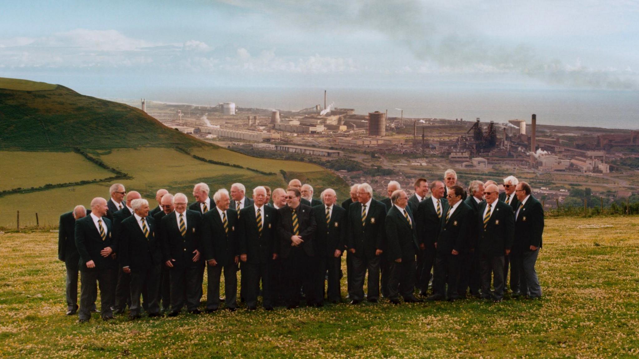 A male voice choir in dark green suits, white shirts and green and yellow striped ties standing on a green hill with yellow daisies. In the background is the industrial town of Port Talbot and the large steelworks. To the left of the frame is another lush green hillside. In the distance is the sea and a blue sky with little cloud and some grey smoke.