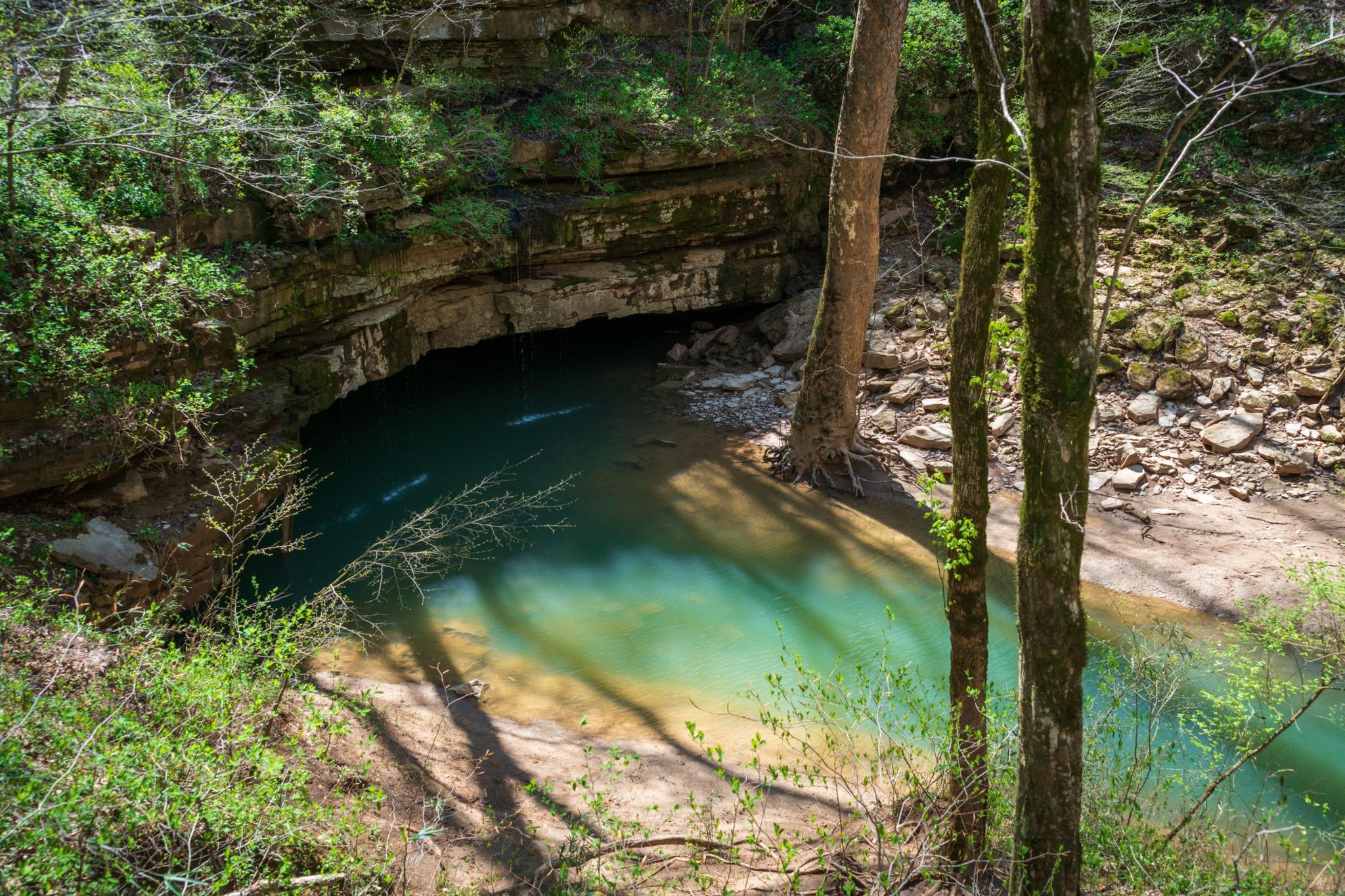a blue-green river with sandy banks and green foliage travelling through trees and into a dark cave