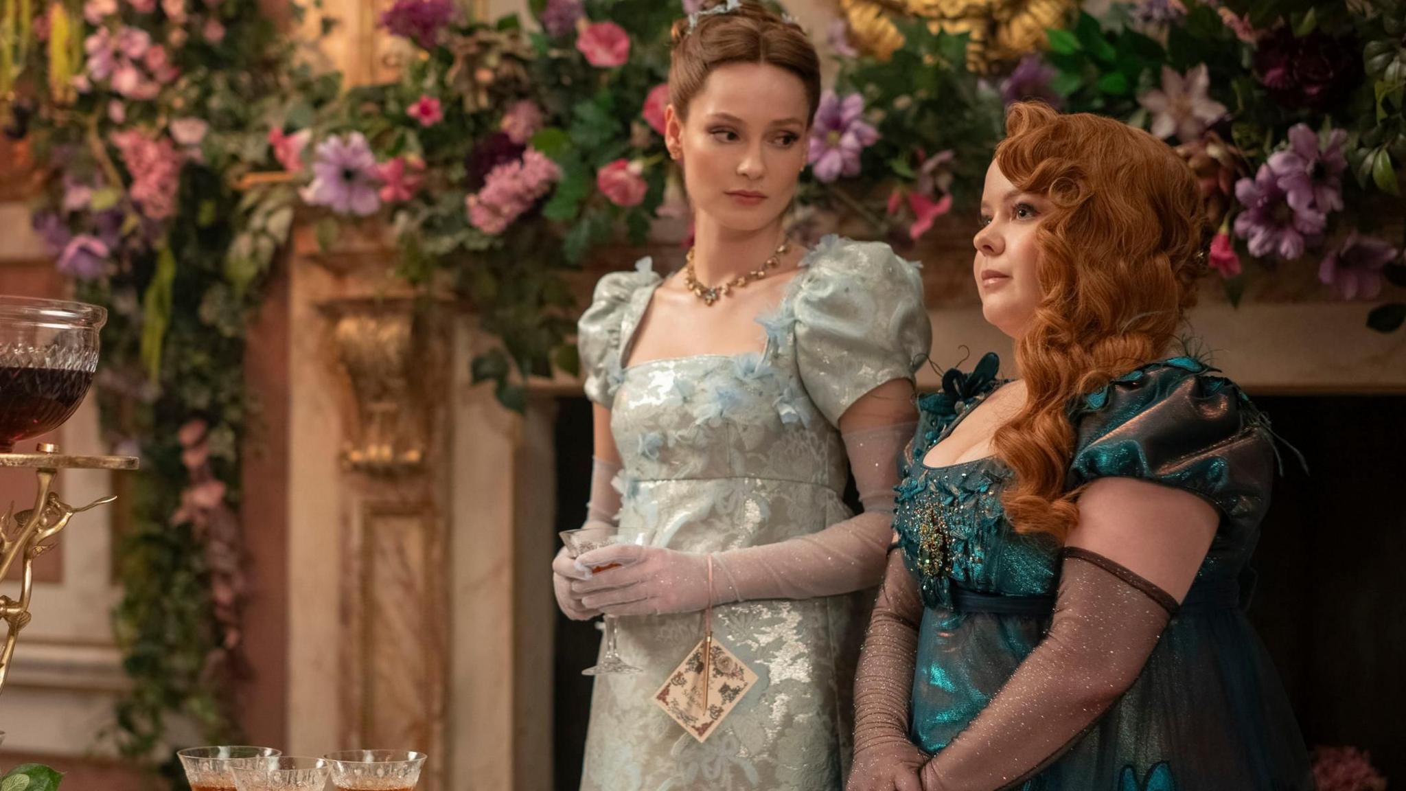 Hannah Dodd (left) as Francesca Bridgerton and Nicola Coughlan as Penelope Featherington in episode 301 of Bridgerton. The women are wearing evening dresses and standing in front of flower decorations.