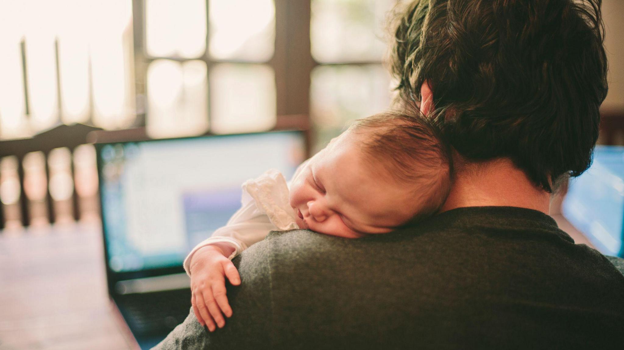 A man holding a baby while working on a laptop. The baby is sleeping on his shoulder.