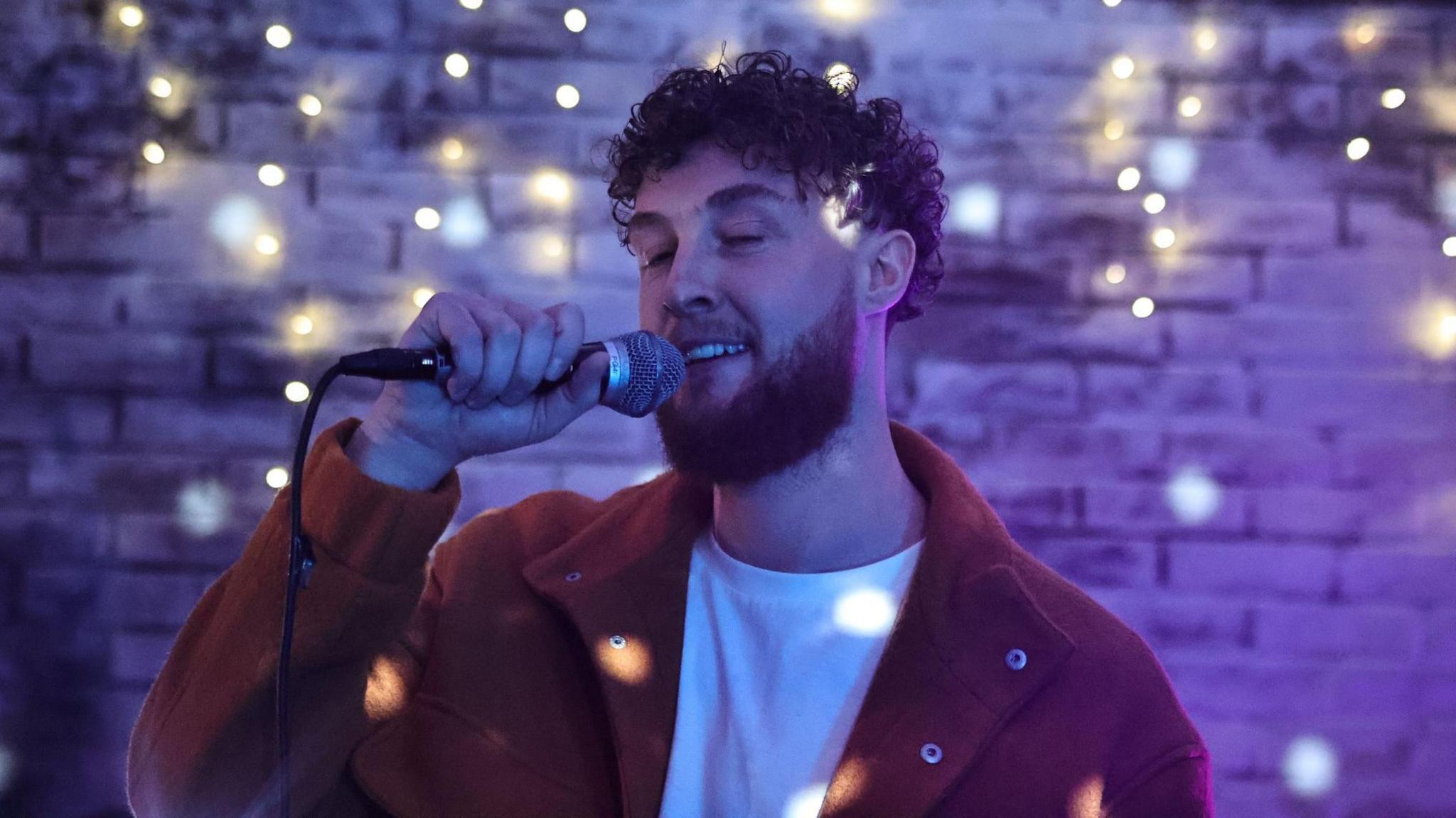 Ryan Finan with brown hair has his eyes half closed as he sings into a microphone against purple lighting under lights reflected of what appears to be a disco ball.