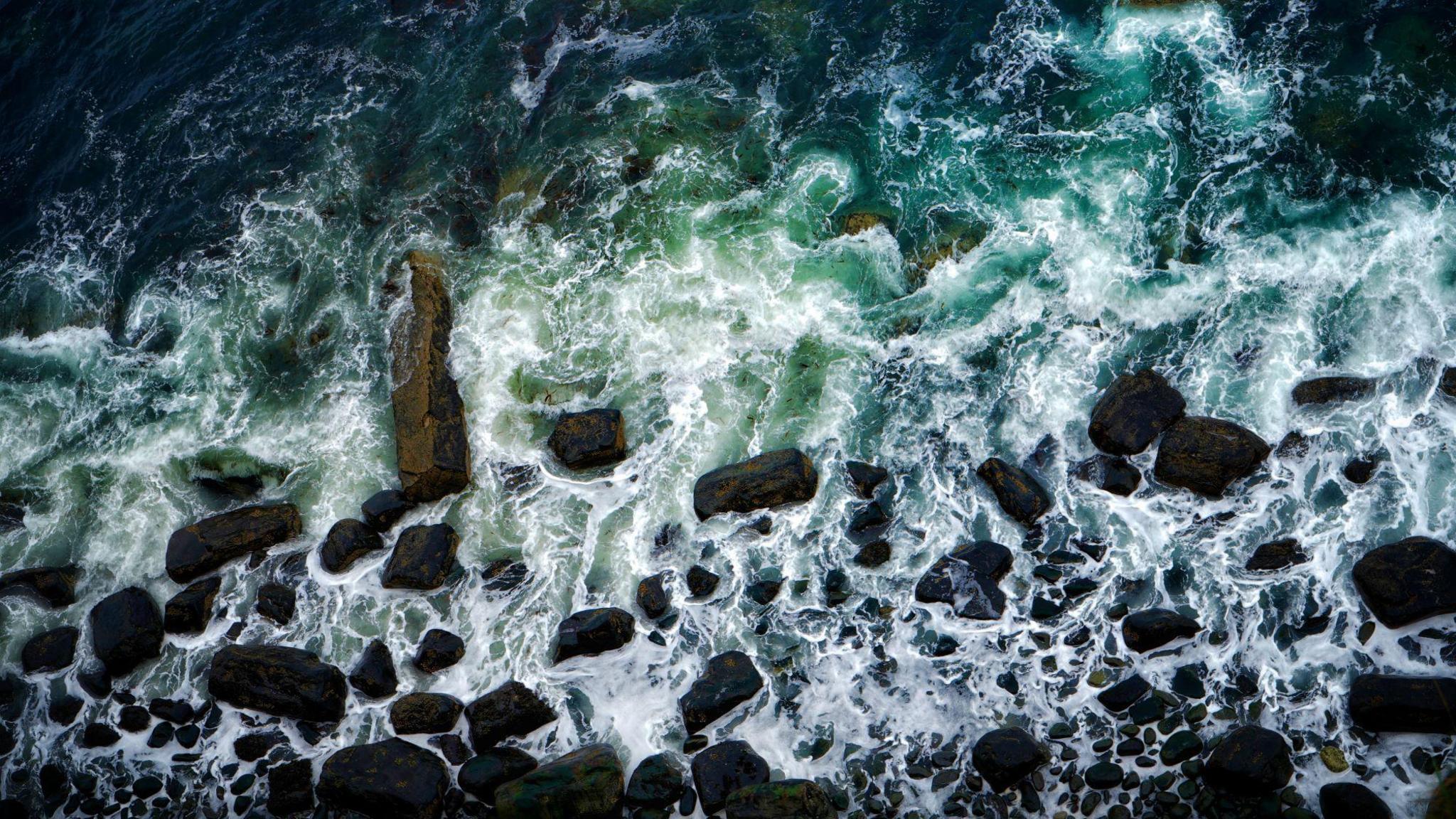 Overhead view of water crashing on rocks, creating white waves near the shoreline 