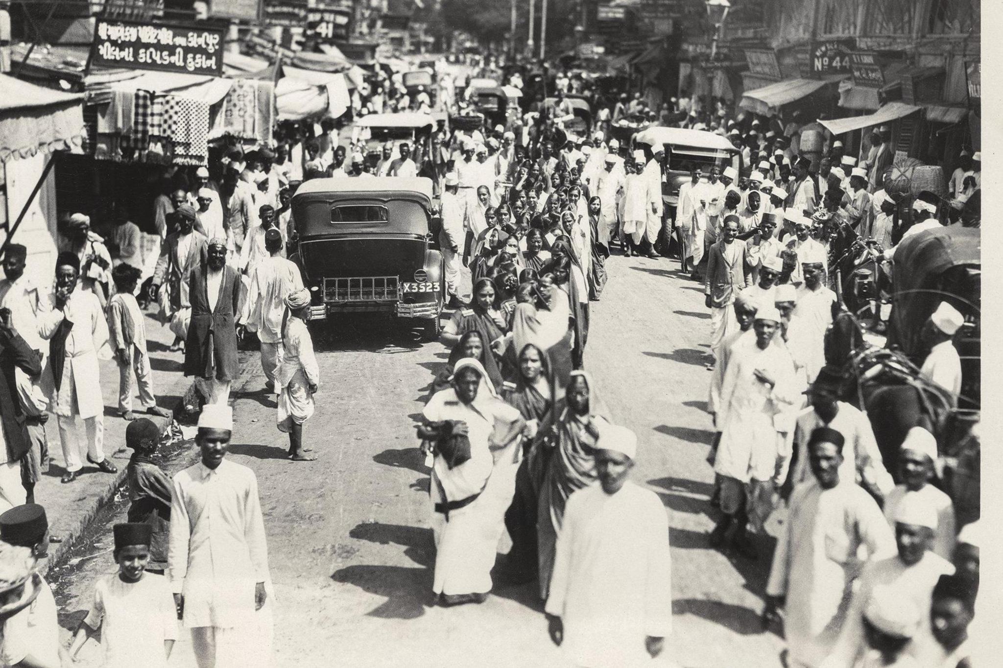 A wide-angle shot of India's bustling streets as men and women, most of whom are protesters, walk through the traffic along a boulevard dotted with shops
