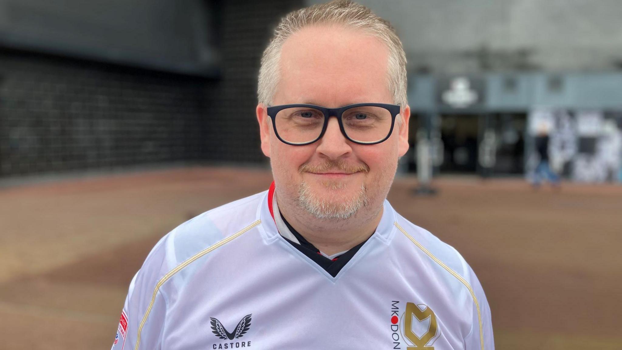 Man in mid 40s with spectacles wears a white football shirt with gold piping. He has a goatee bears and stands outside a football stadium.