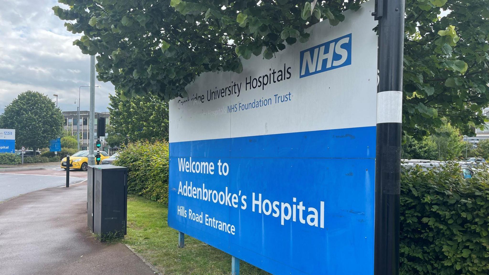 Exterior of Addenboroke's Hospital. There is a blue and white sign and a yellow car waiting at traffic lights.