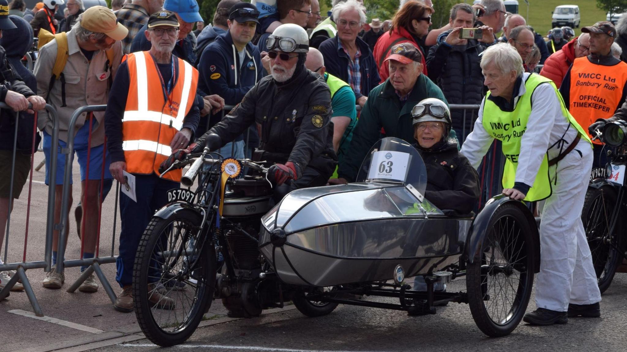 A man with a white beard, wearing black clothing, sunglasses and a helmet with motorcycle goggles on it, sits on a vintage motorcycle. Crowds of people are watching from the other side of a barrier. A woman wearing similar gear sits in a sidecar. Crowds of people are watching from the other side of a barrier and a man in a hi-viz tabard is leaning on the back of the vehicle.