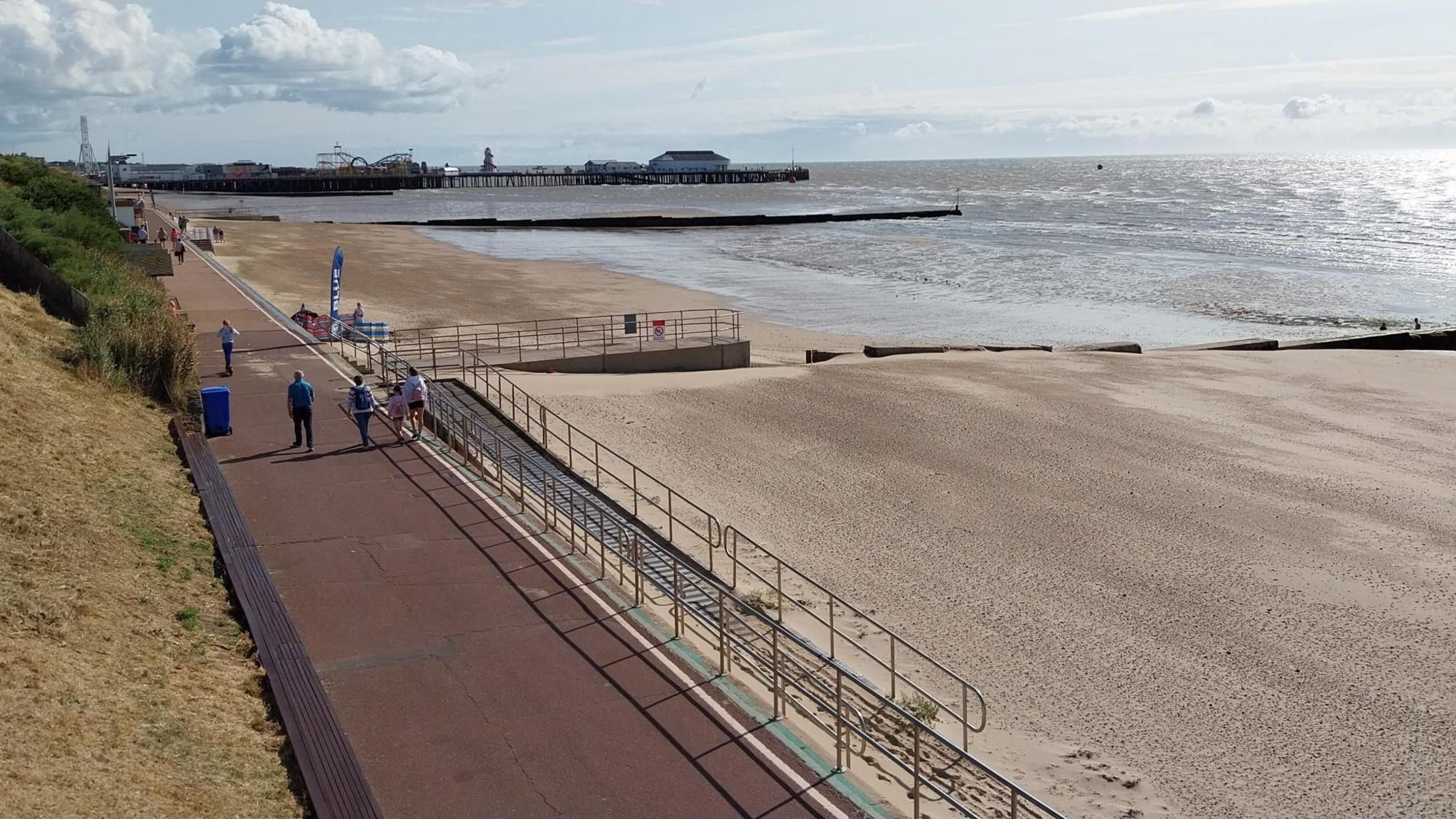 An empty beach has railings along the seafront and a brown path leading into the distance. Clacton Pier is in the far distance with the silhouette of a roller coaster and helter skelter. The tide is out and the sea is shining to the right of the photo. The sky is light in colour with some clouds.