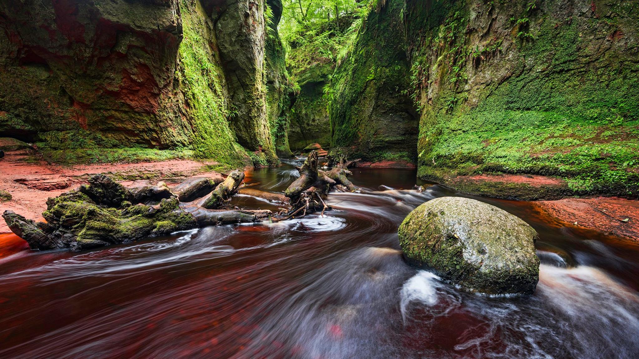 A long exposure shot of water flowing down a steep gorge. A round rock can be seen in the foreground and steep green covered rocks can be seen in the background